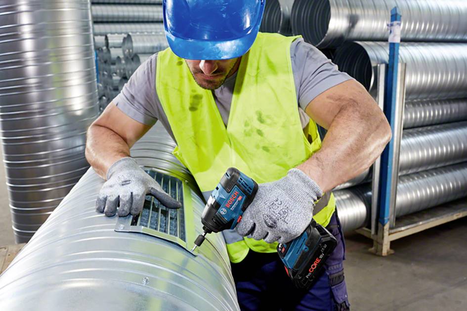 Ein Arbeiter mit blauer Schutzkappe und gelber Weste schraubt mit einem Akkuschrauber an einem großen Lüftungsrohr in einer Werkstatt.