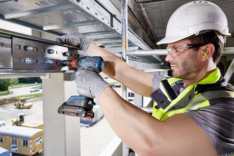 Ein Bauarbeiter mit Schutzhelm und Handschuhen montiert Metallträger mit einer elektrischen Bohrmaschine auf einer Baustelle.