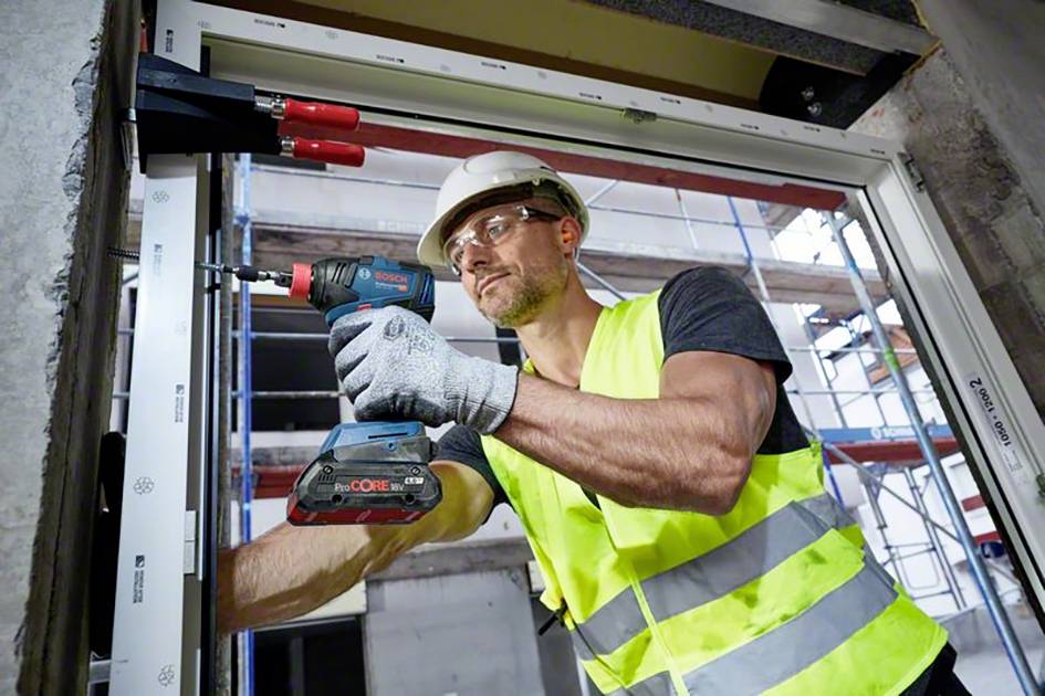 Ein Bauarbeiter mit Helm und Warnweste befestigt ein Fensterrahmen mit einer elektrischen Bohrmaschine auf einer Baustelle.