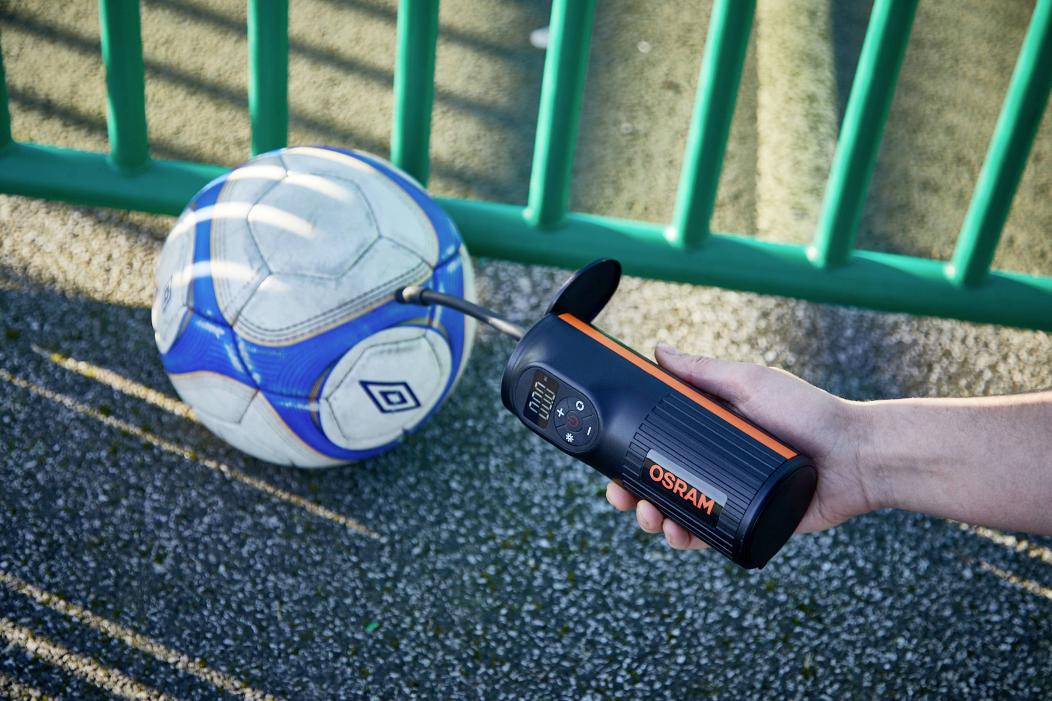 Eine Person pumpt einen Fußball mit einem tragbaren Gerät auf, während der Ball auf einer gepflasterten Oberfläche neben einem grünen Zaun liegt.