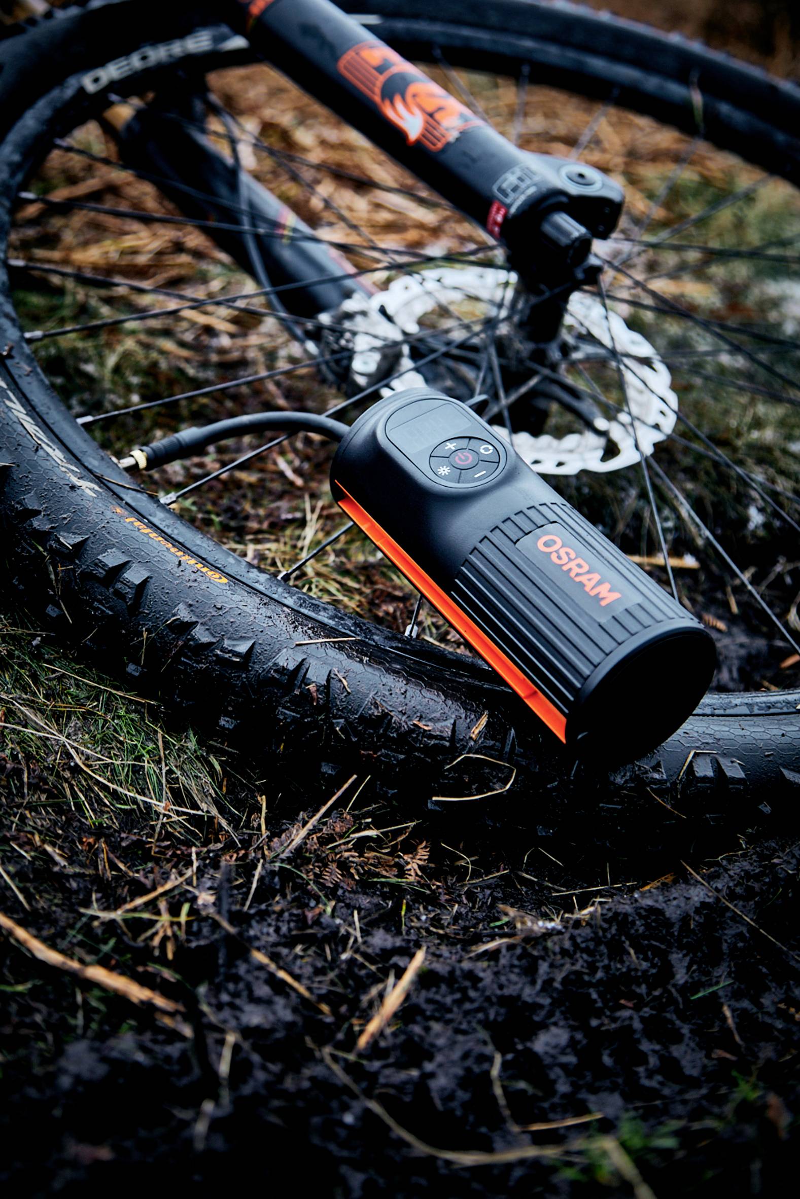 Fahrradreifen mit Druckluftpumpe in feuchter Umgebung. Ein Mountainbike liegt auf dem Boden, die Pumpe ist markiert mit 'OSRAM'.