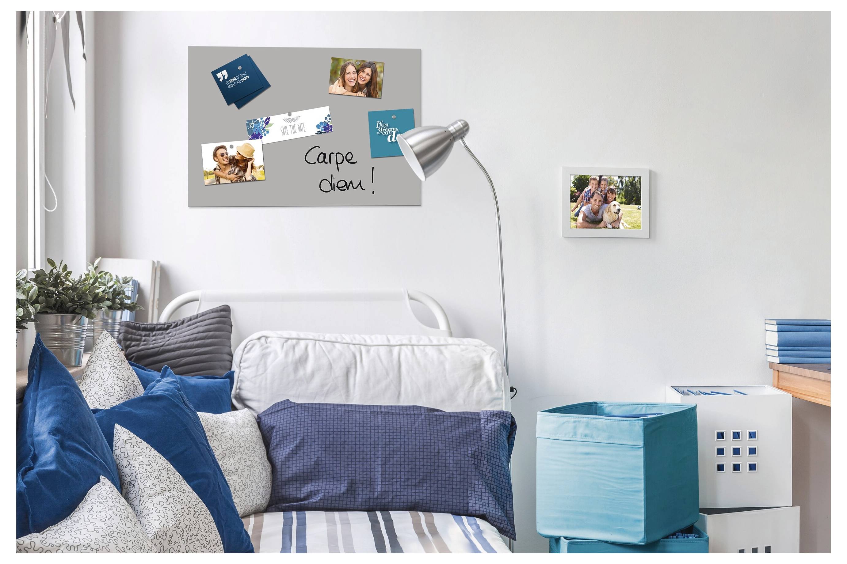 Ein ordentlich gemachtes Bett mit blauen Kissen steht in einem hellen Schlafzimmer neben einer silbernen Stehleuchte, einer Wandtafel mit Fotos und „Carpe diem!" sowie Aufbewahrungsbehältern.