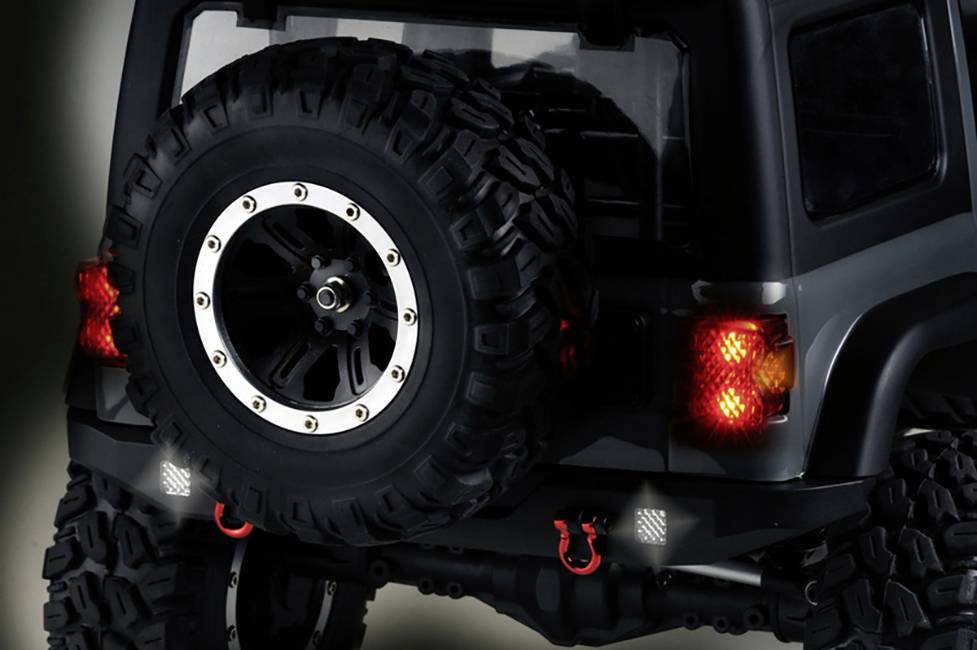 Ein Geländewagen mit großen Reifen und zwei roten Haken hinten. Rücklichter leuchten. Das Auto scheint für Offroad-Abenteuer bereit zu sein.