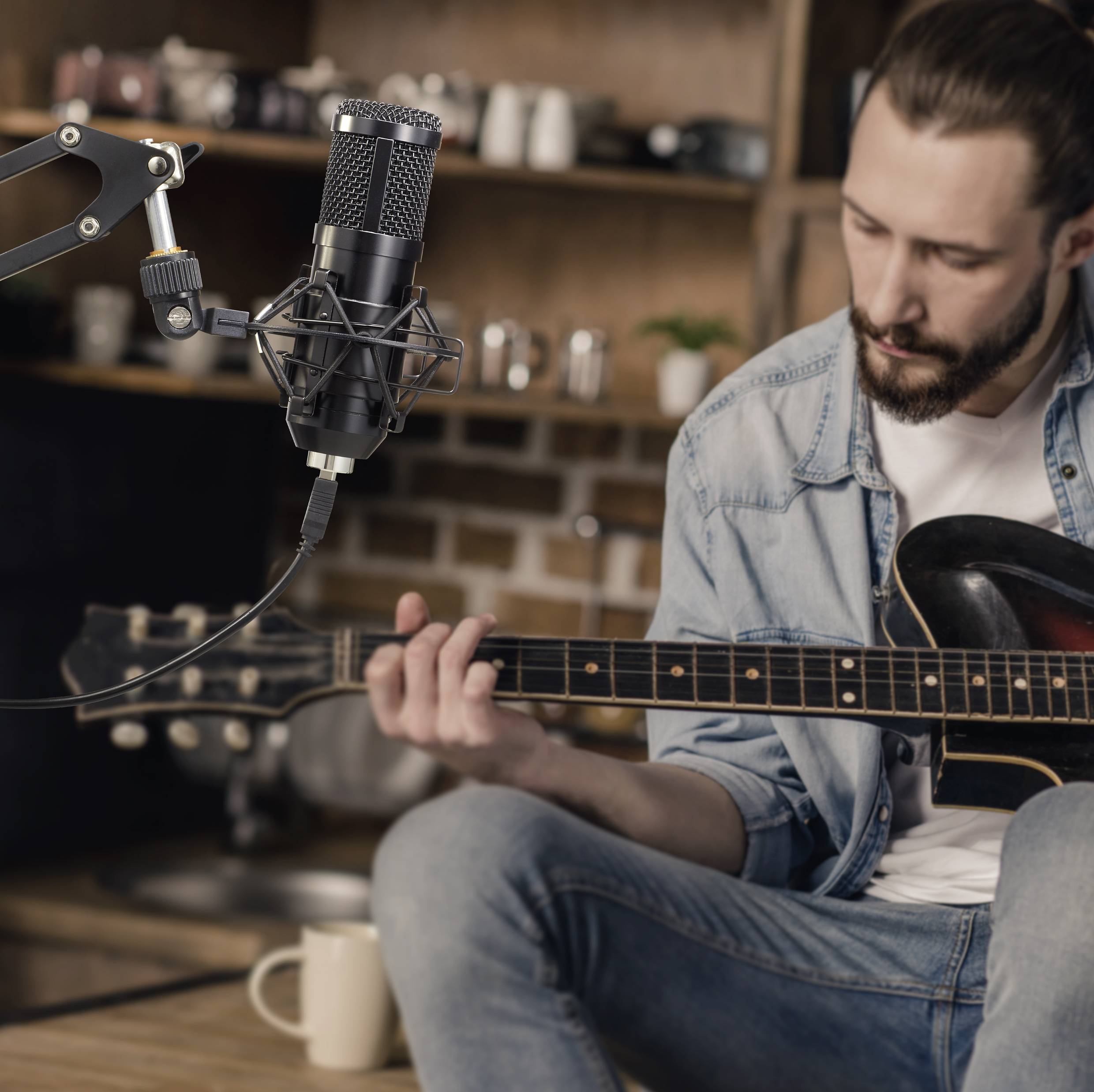 Ein Mann spielt Gitarre in einem gemütlichen Raum. Ein Mikrofon ist auf ihn gerichtet, wahrscheinlich für eine Aufnahme oder ein Streaming.