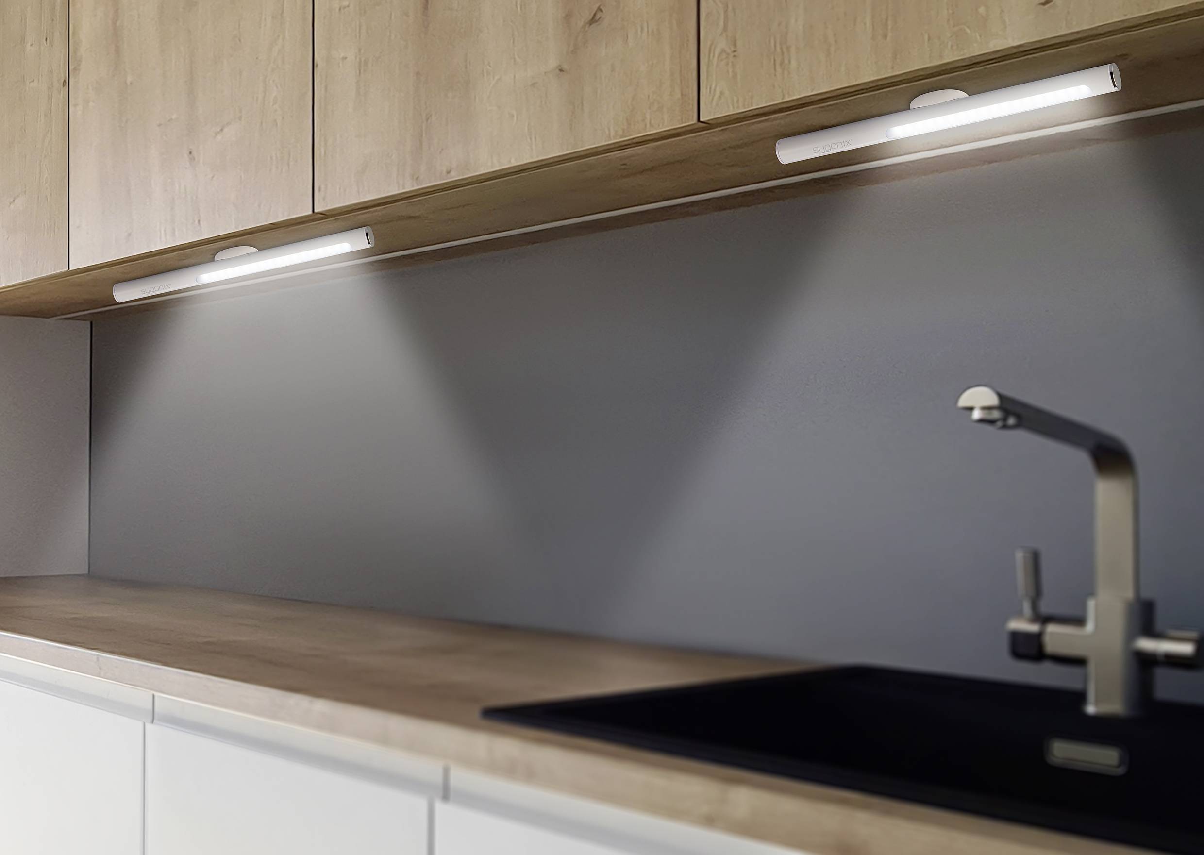 Innenküche mit modernen Holzschränken, beleuchtet durch zwei Unterbauleuchten, graue Rückwand und Edelstahlarmatur über schwarzem Spülbecken.