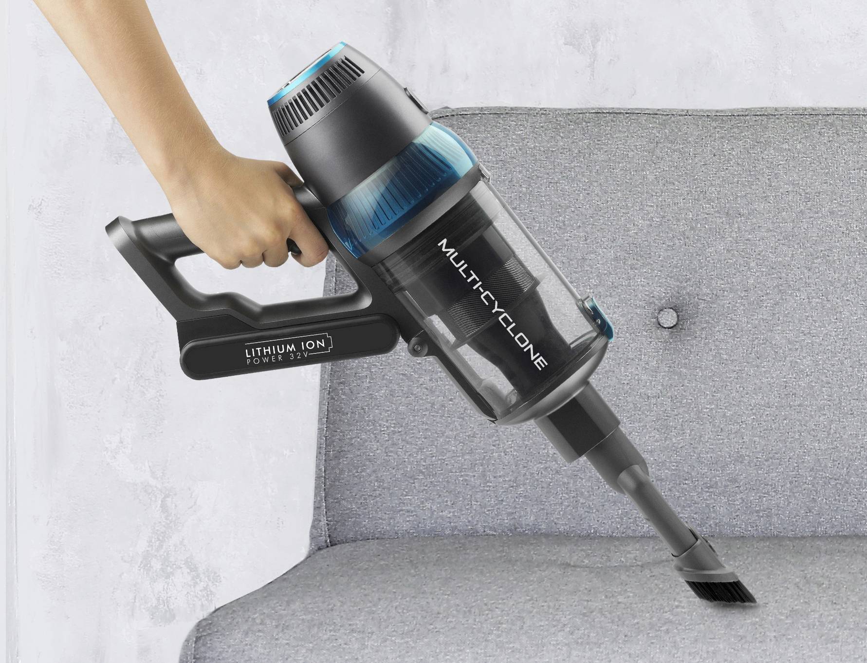 Person hält einen kabellosen Handstaubsauger und reinigt ein graues Sofa. Staubsauger mit blauer Akzentfarbe, markiert mit 'Multicyclone'.