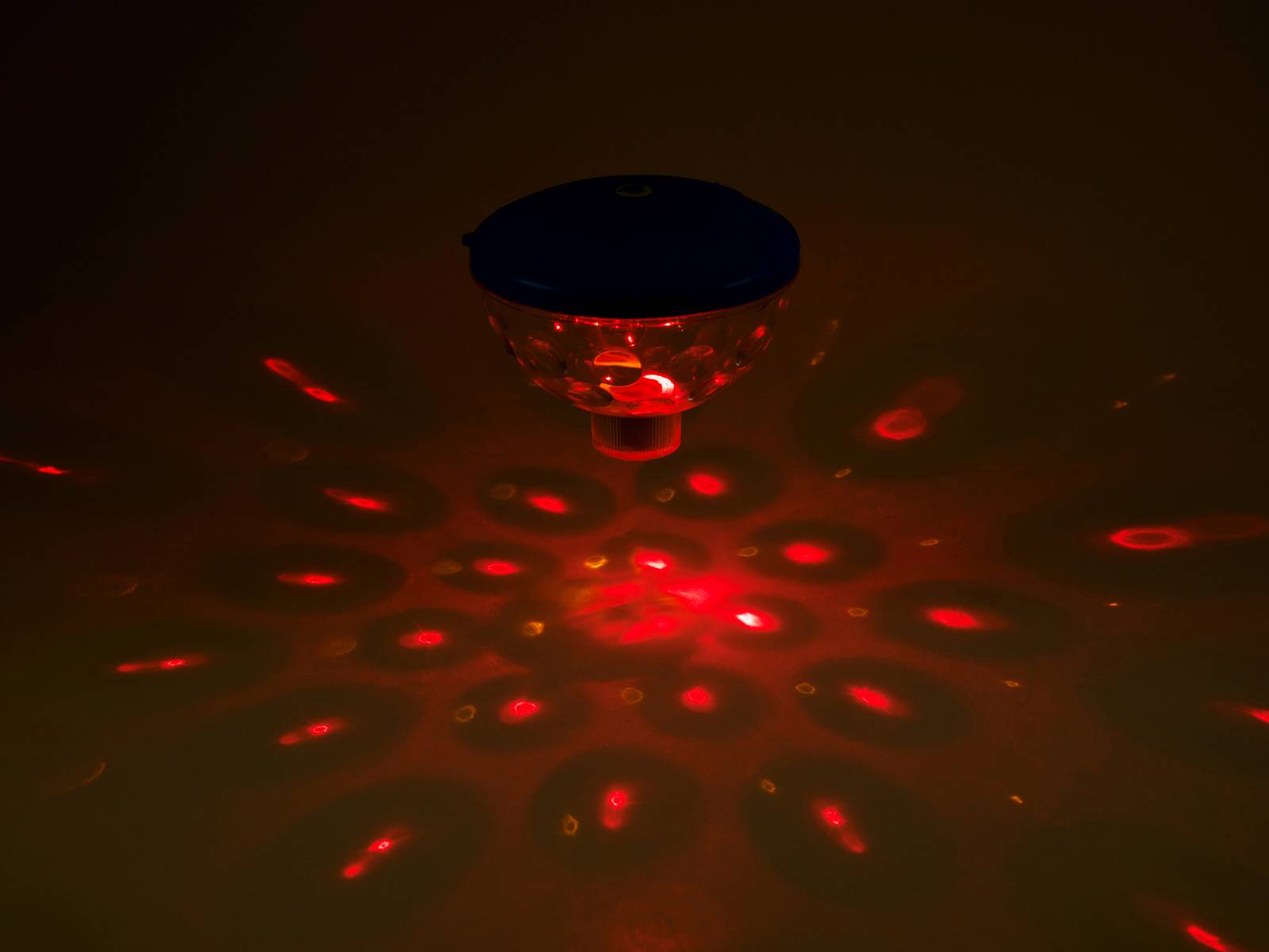 Ein rot beleuchtetes Gerät projiziert ein kreisförmiges Muster aus roten Punkten und Ovalen auf eine dunkle Oberfläche.