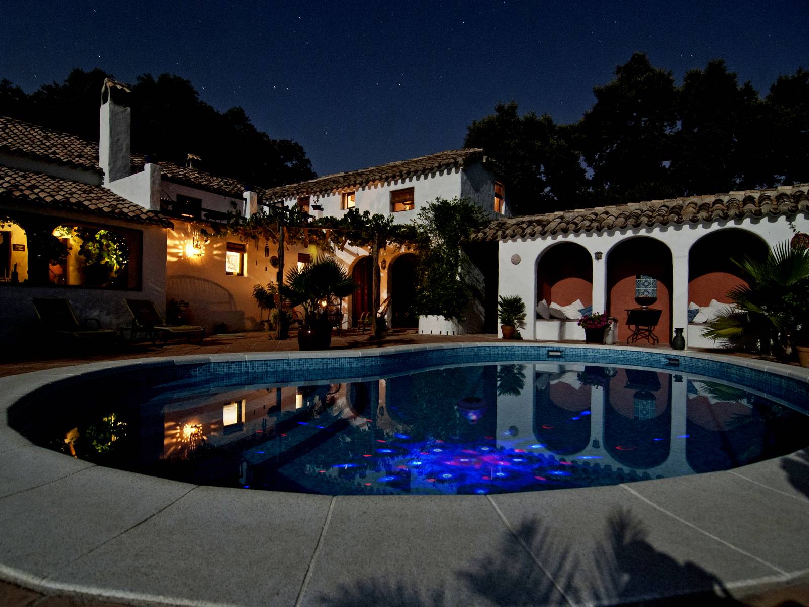 Ein beleuchteter Pool bei Nacht vor einem großen, spanischen Haus mit Terrakottadach. Lichter spiegeln sich im Wasser.