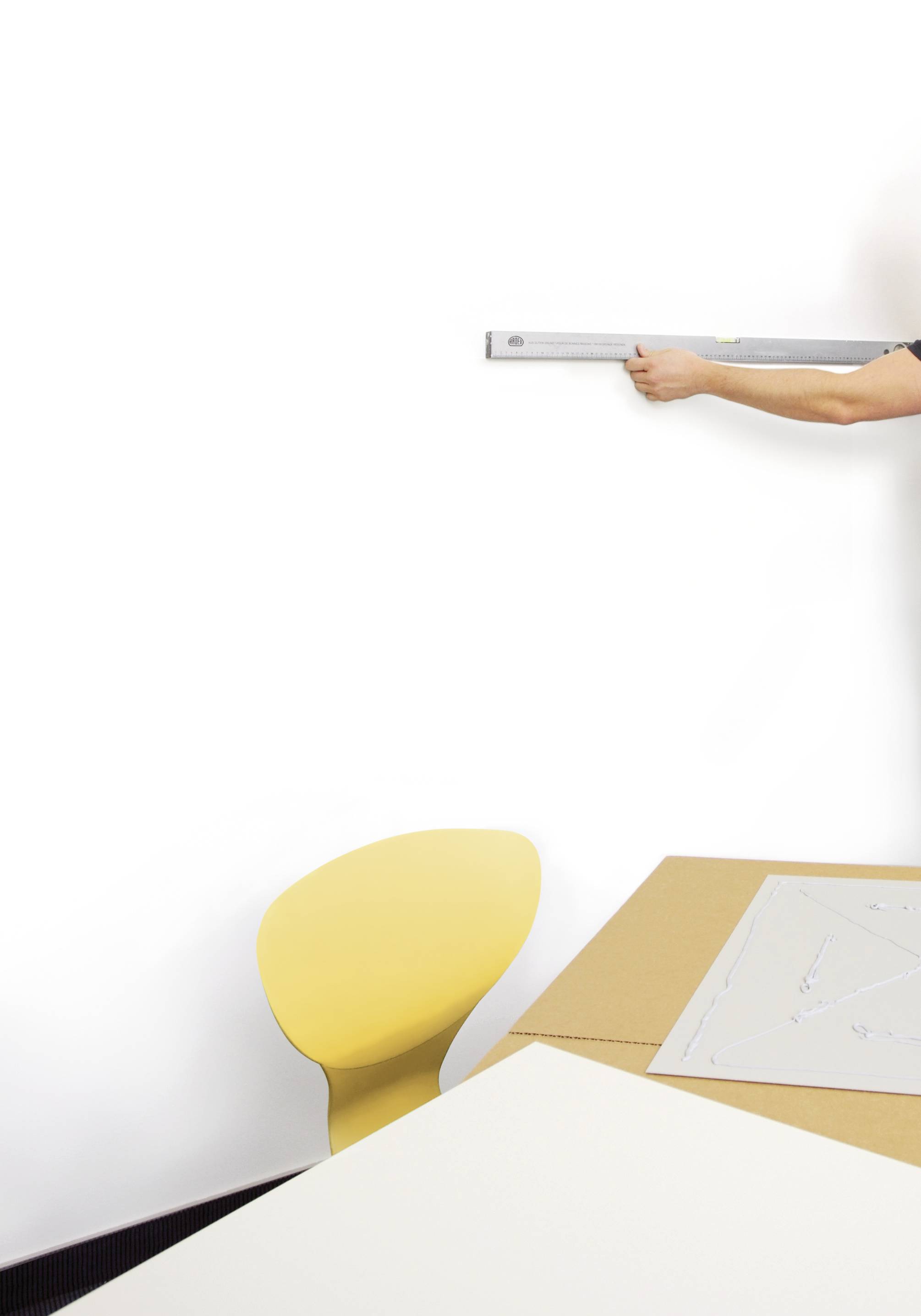 Person misst eine leere weiße Wand mit einem Lineal, gelber Stuhl und Tisch im Vordergrund, Vorbereitung für Dekoration oder Bau.
