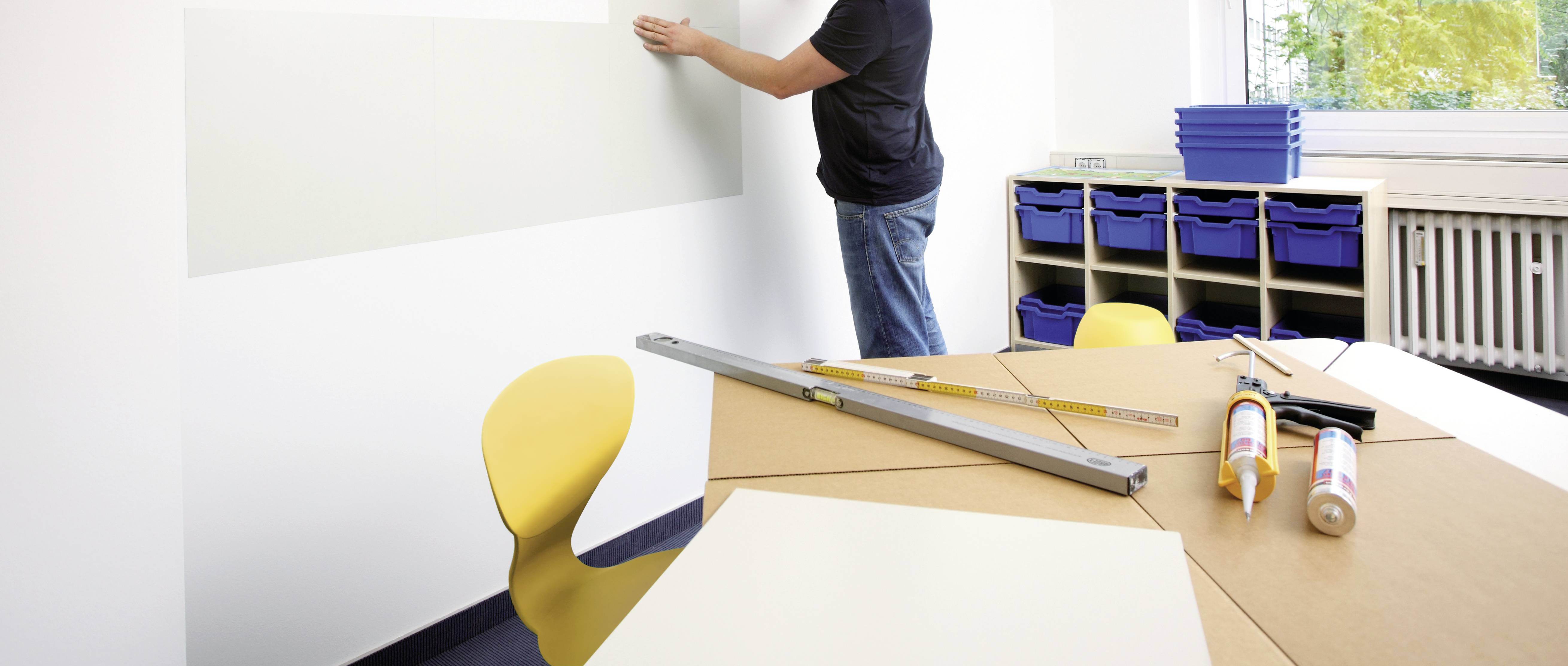 Eine Person montiert ein hellgraues Whiteboard an einer weißen Wand. Im Vordergrund ein Tisch mit Werkzeugen und Klebstoff.