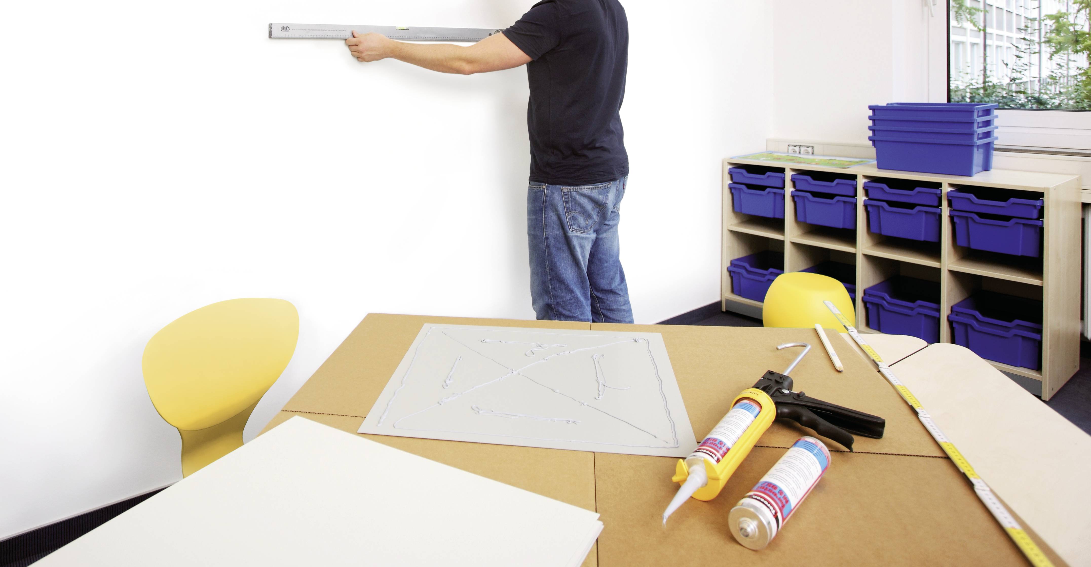 Eine Person misst eine weiße Wand mit einer Wasserwaage aus. Im Vordergrund liegen Werkzeuge und Baumaterialien auf einem Tisch.