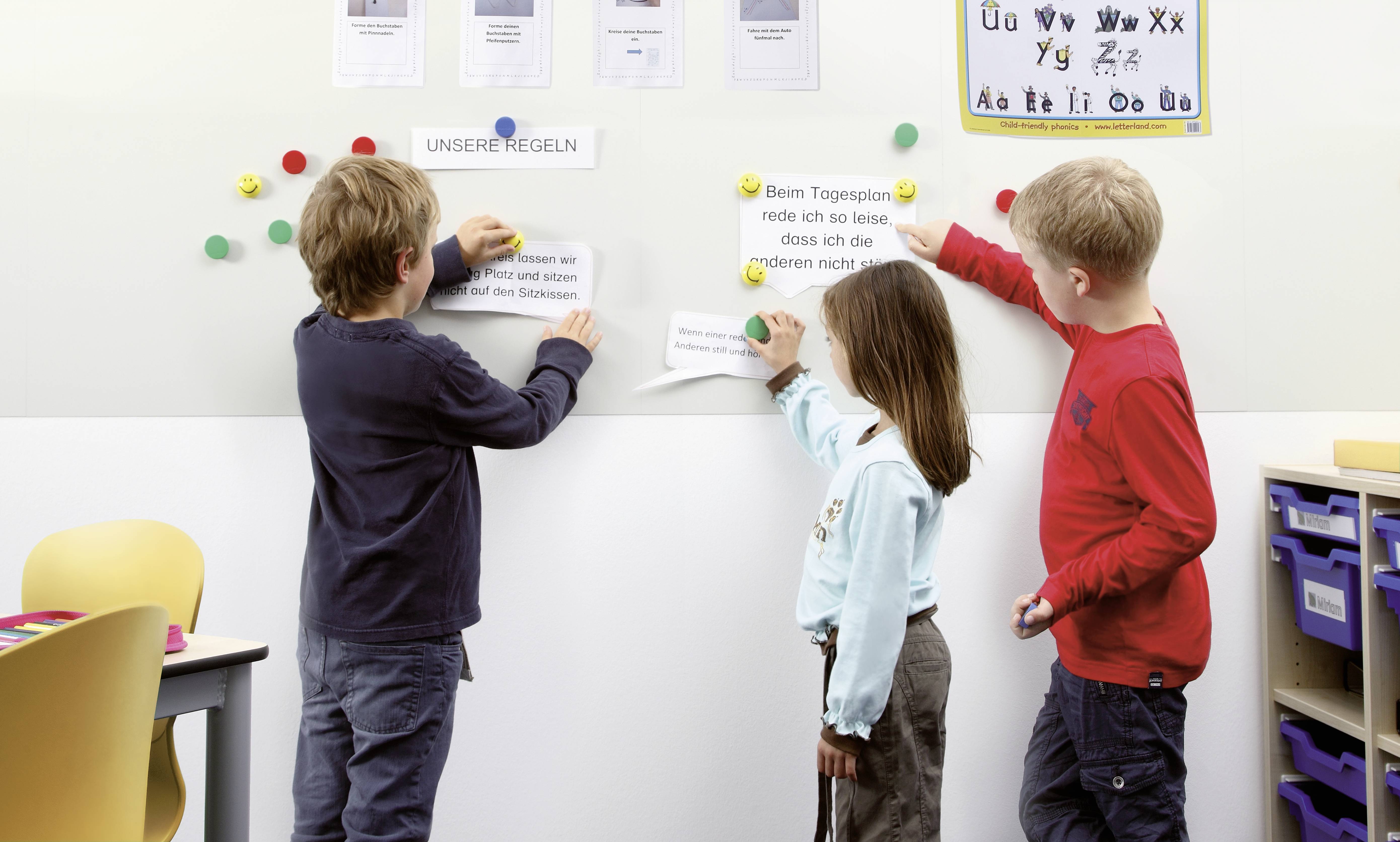 Drei Kinder hängen Papier mit Regeln an eine Wand in einem Klassenzimmer. Im Hintergrund sind Schilder mit Buchstaben und mehr Regeln sichtbar.