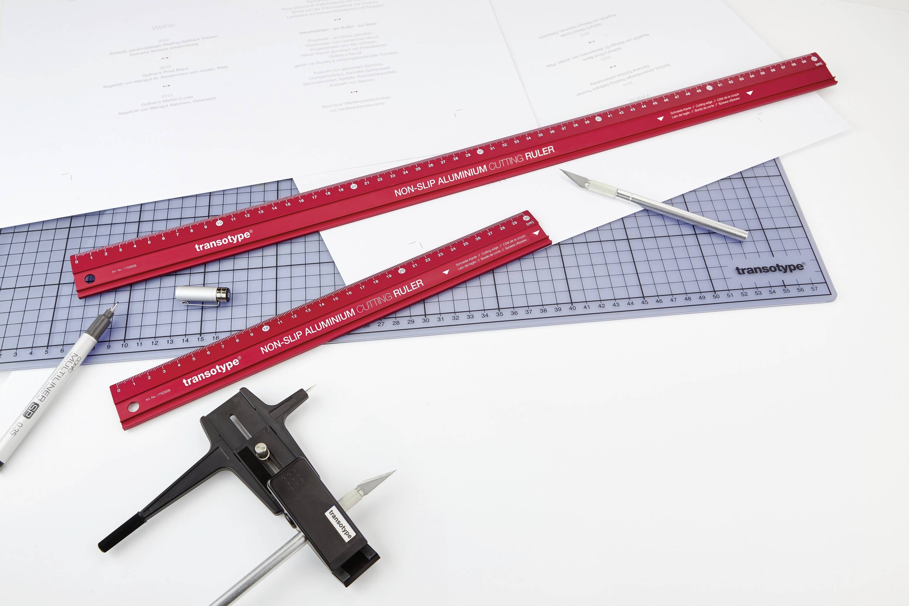 Zwei rote Lineale, ein Stift und weiteres Zeichenzubehör liegen auf einem Zeichenbrett. Hintergrund zeigt Entwürfe und Linealmarkierungen.