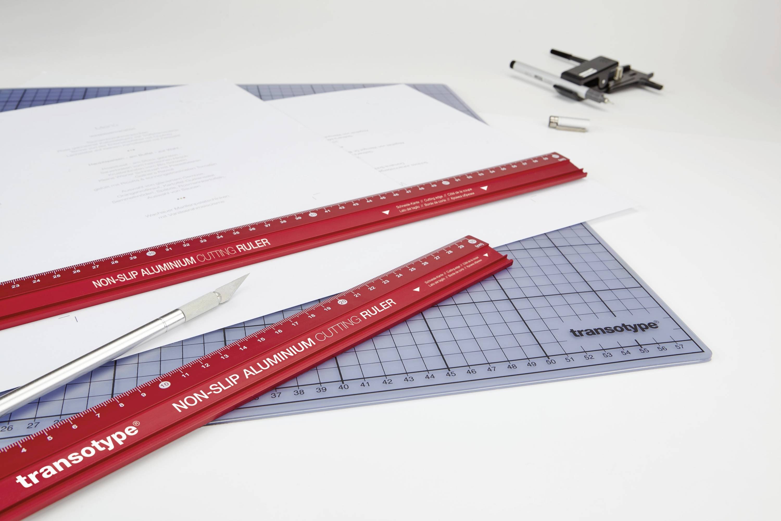 Roter Aluminiumlineal mit der Aufschrift 'NON-SLIP ALUMINIUM CUTTING RULER' liegt auf einem Schneidebrett. Daneben ein Cutter und Stifte.
