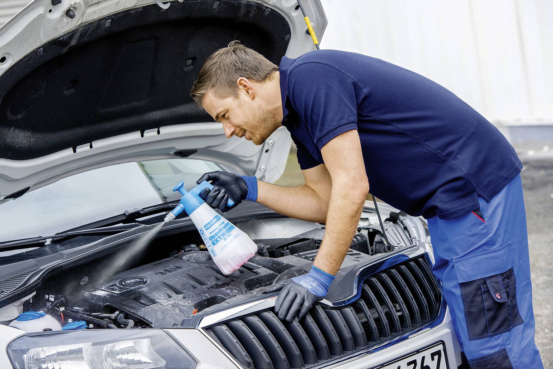 Ein Mechaniker reinigt den Motorraum eines Autos mit einem Sprühreiniger. Der Autohimmel ist offen, der Mann trägt Arbeitshandschuhe.