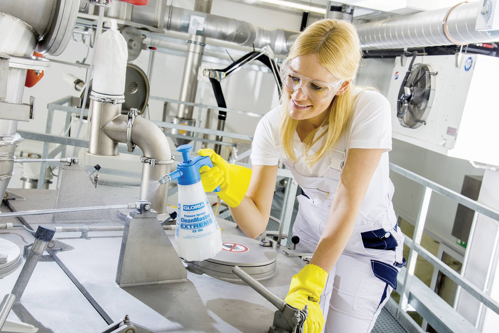 Eine Frau in Schutzkleidung und Handschuhen reinigt industrielle Maschinen mit einem Sprühmittel in einer Fabrikumgebung.