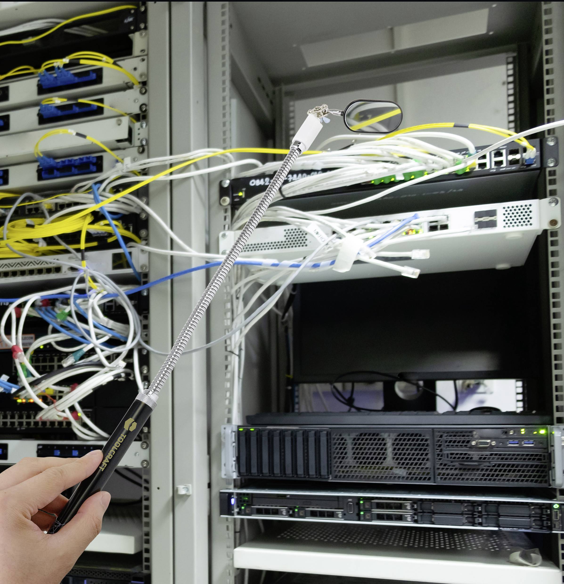 Eine Hand hält einen Teleskopspiegel vor einem Server-Rack mit Netzwerkkabeln, um schwer zugängliche Bereiche zu inspizieren.