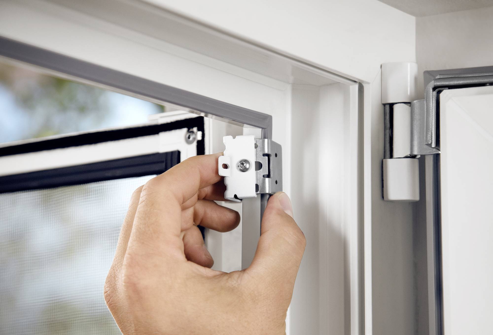 Eine Hand montiert ein kleines, weißes Metallteil an einem Fensterrahmen. Im Hintergrund sind ein geöffnetes Fenster und eine Fliegengittertür zu sehen.