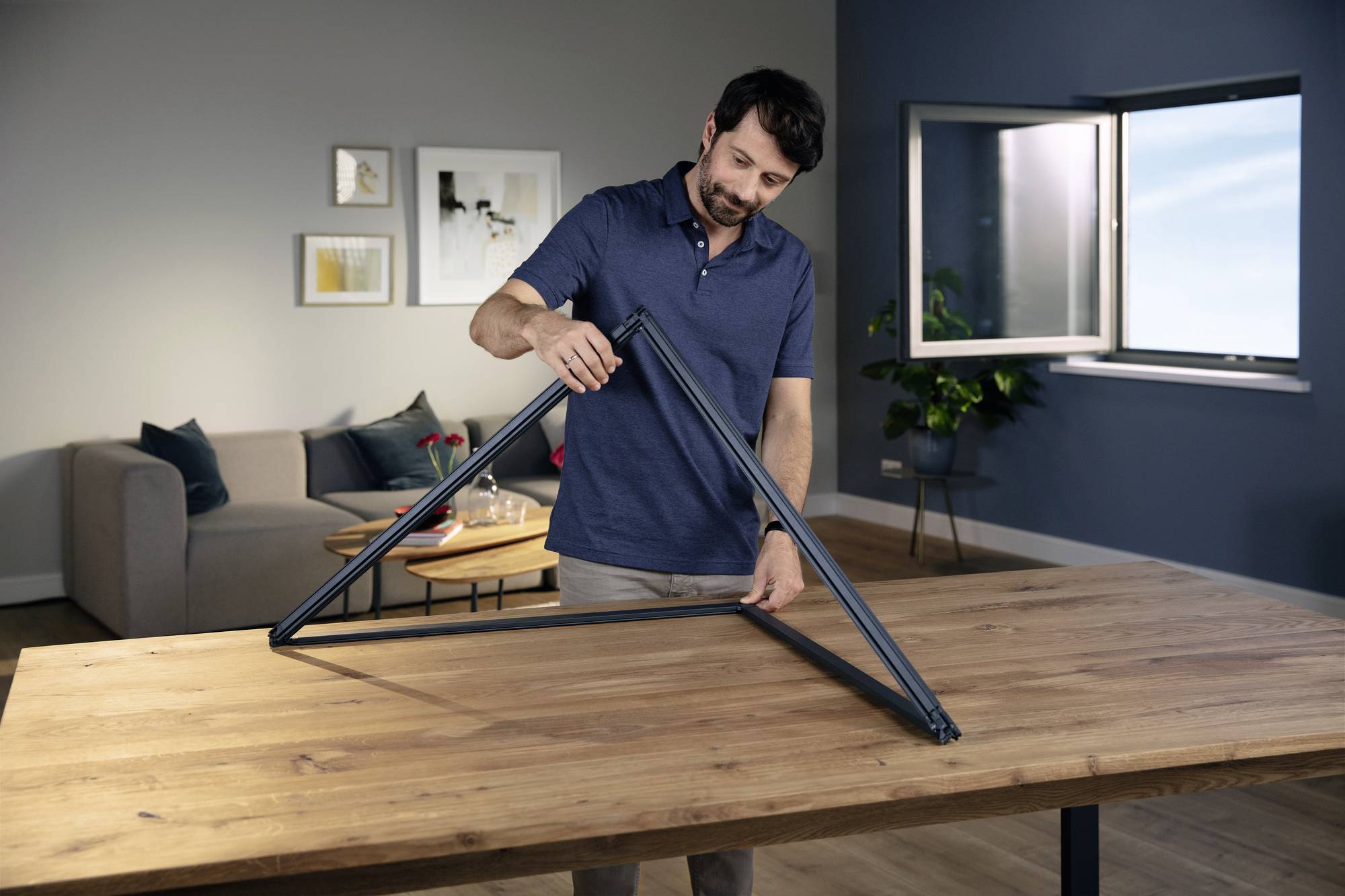 Ein Mann montiert einen Metallrahmen auf einem Tisch in einem modern eingerichteten Wohnzimmer. Ein Fenster ist geöffnet.