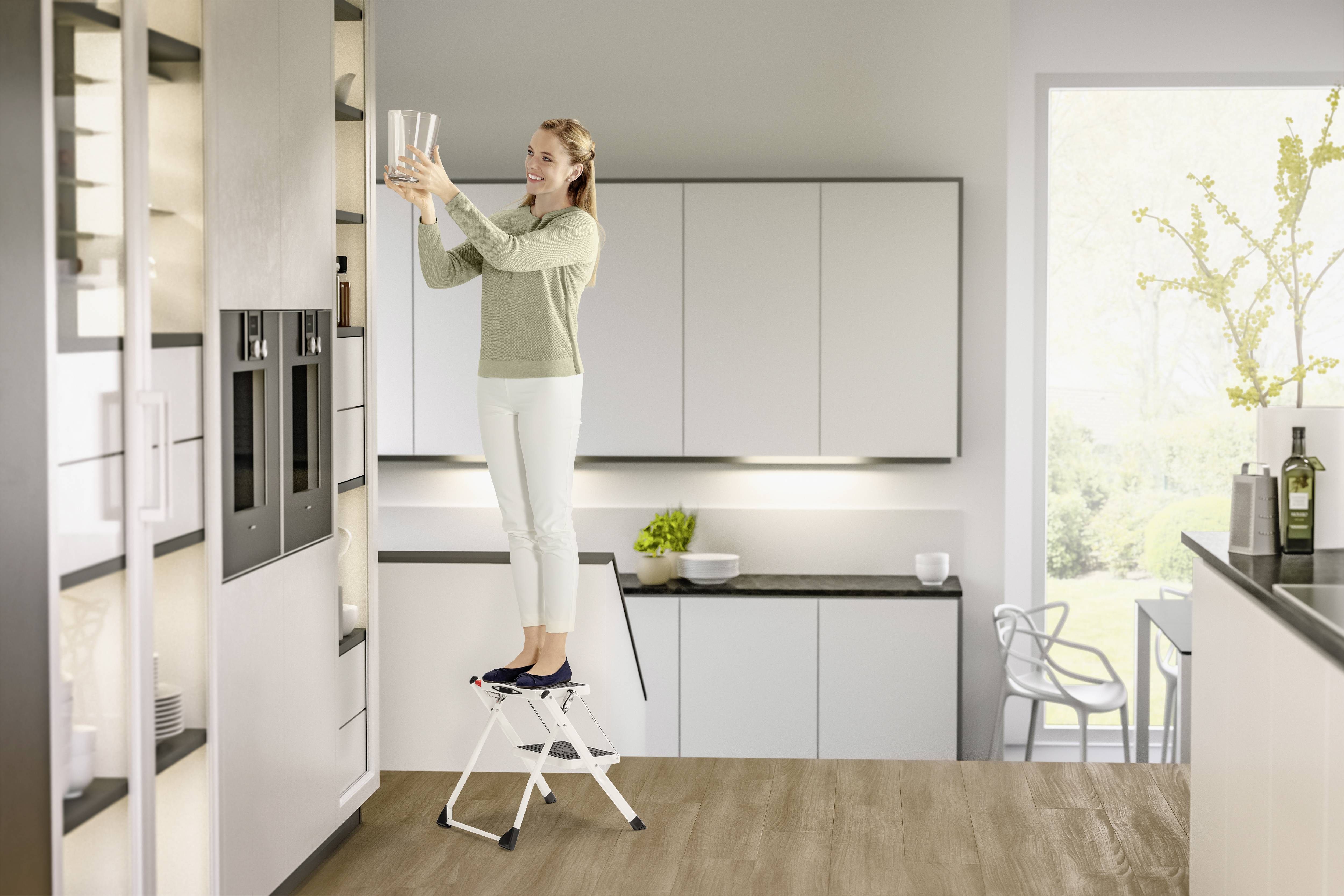 Frau steht auf einem Tritthocker und stellt Gläser in ein oberes Regal einer modernen Küche. Sie trägt helle Kleidung.