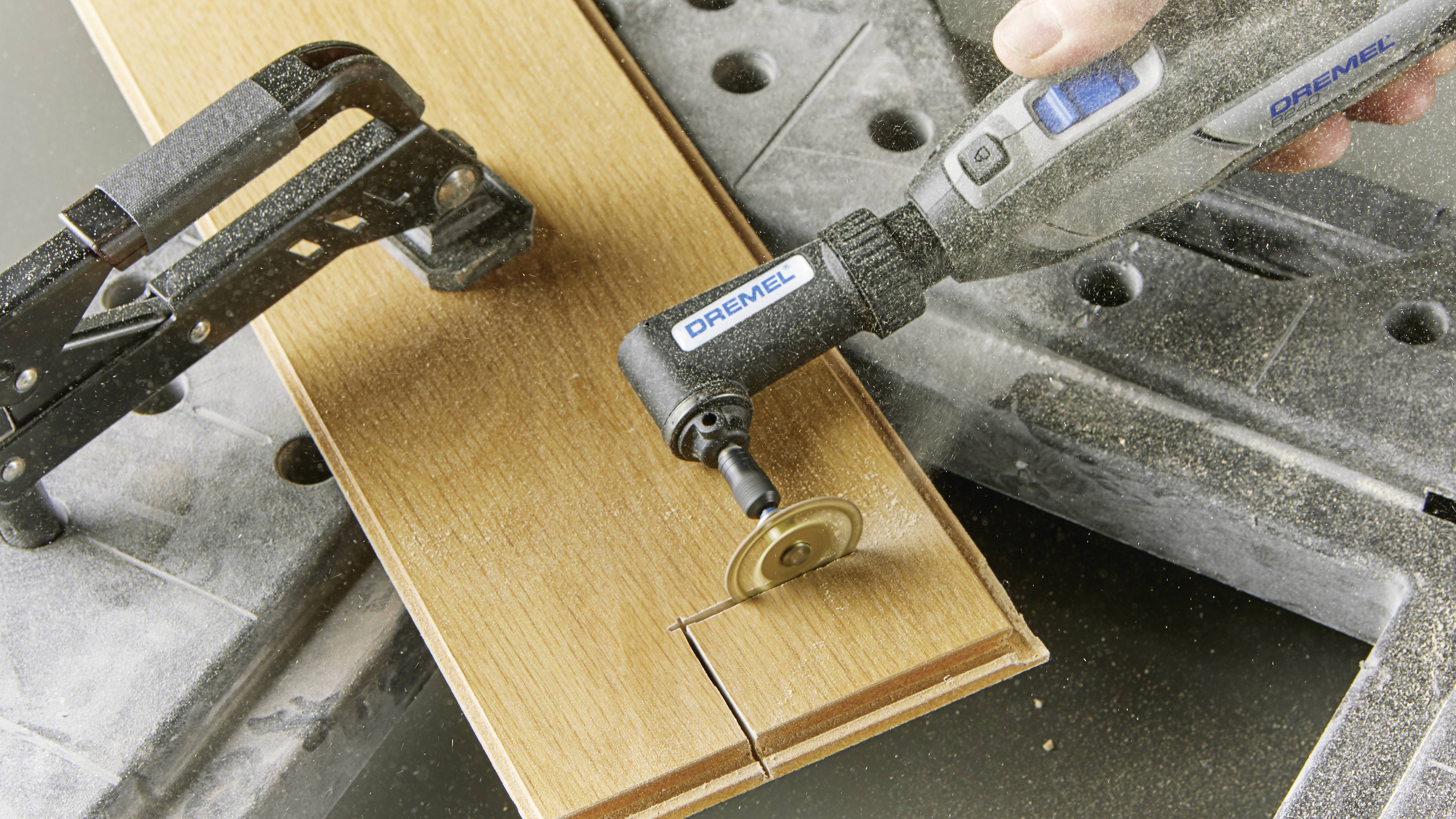 Eine elektrische Handfräse schneidet ein Holzbrett, das mit einer Klammer fixiert ist. Staubpartikel erheben sich während des Schneidens.