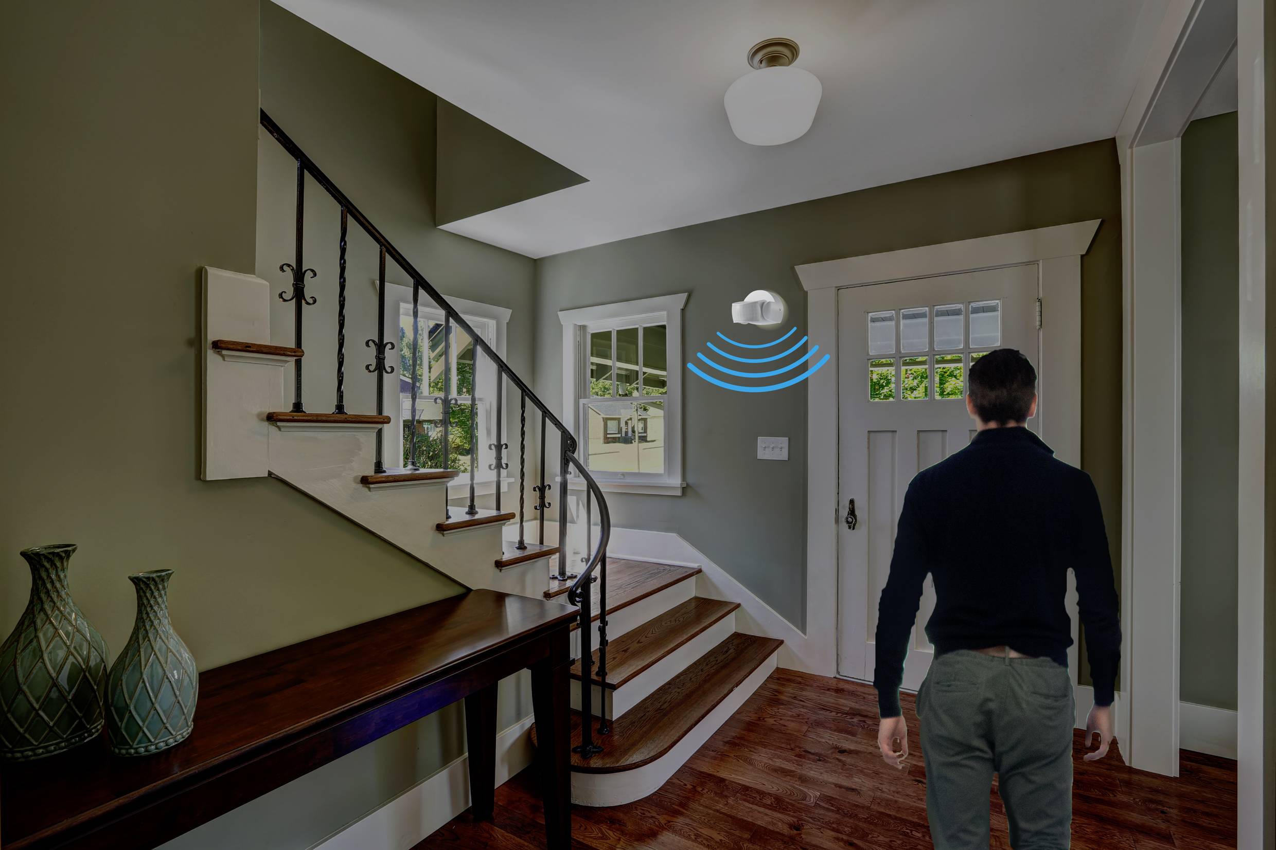 Ein Mann betritt einen Flur mit einem Treppenaufgang. An der Decke ist ein Bewegungsmelder angebracht, der aktiv ist.