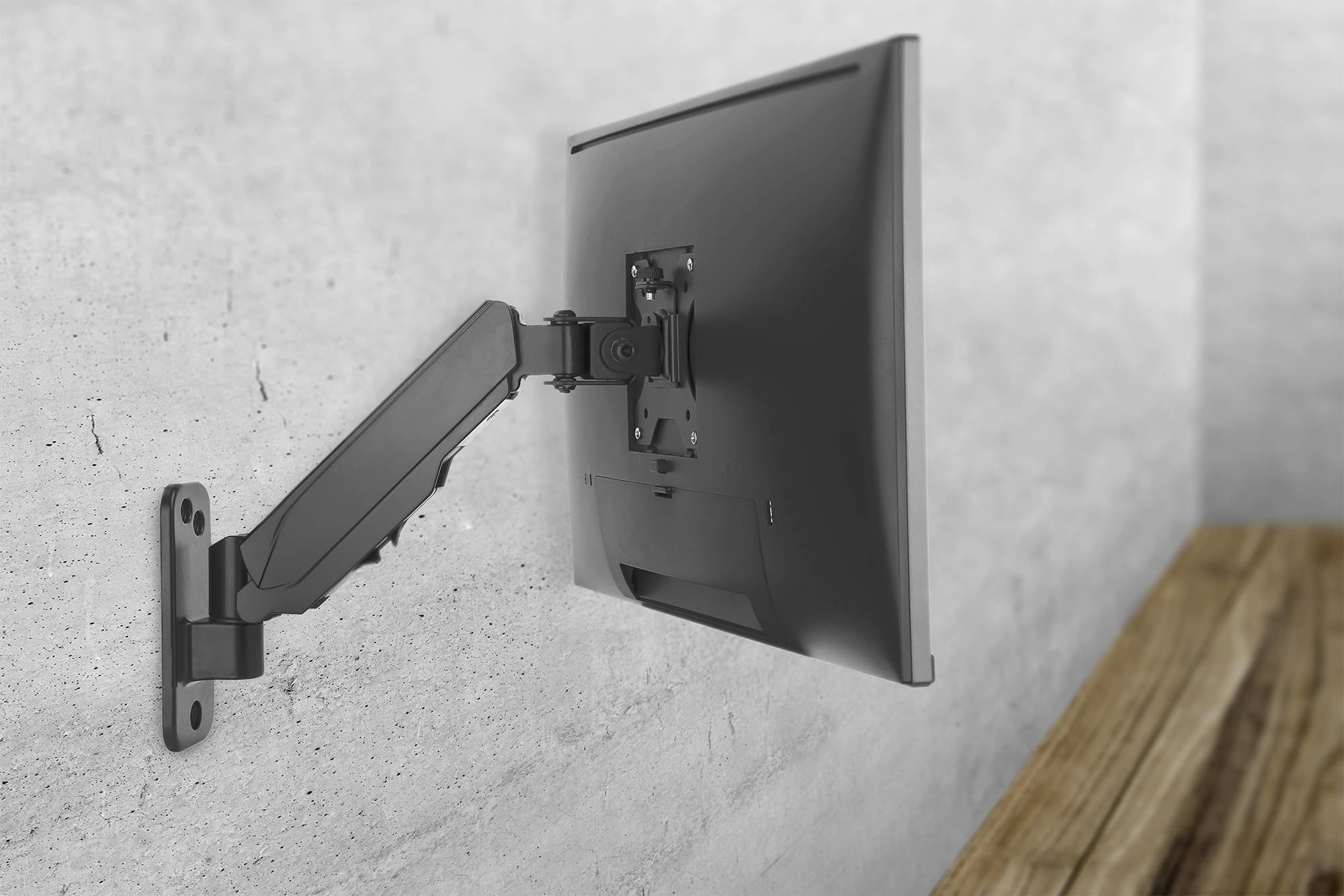 Ein schwarzer Computermonitor ist an einem beweglichen Arm an einer grauen Wand montiert. Der Schreibtisch aus Holz steht daneben.