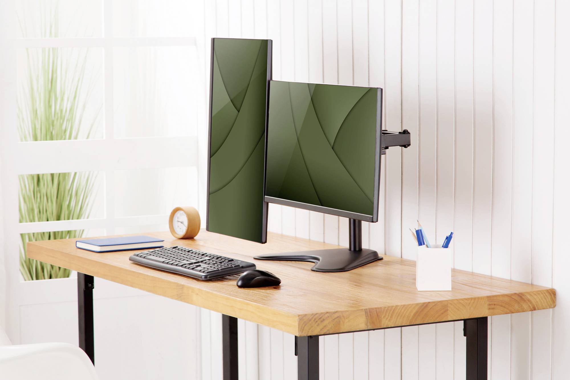 Ein Schreibtisch mit zwei Monitoren, einem Keyboard, einer Maus, einem Notizblock und einem Stifthalter, vor weißer Wand.