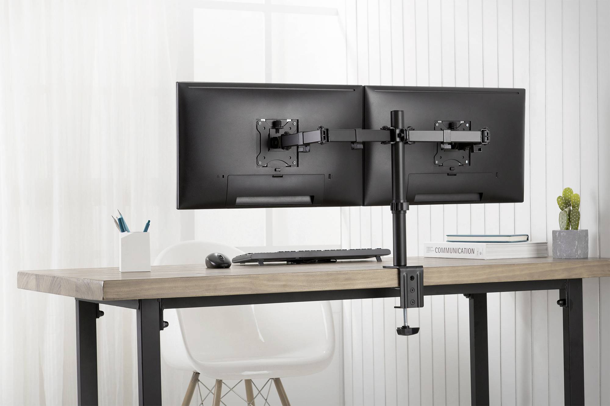 Zwei Monitore auf einem Schreibtisch mit Dual-Monitor-Halterung; im Hintergrund Fenster, daneben Stuhl und Dekorationen.