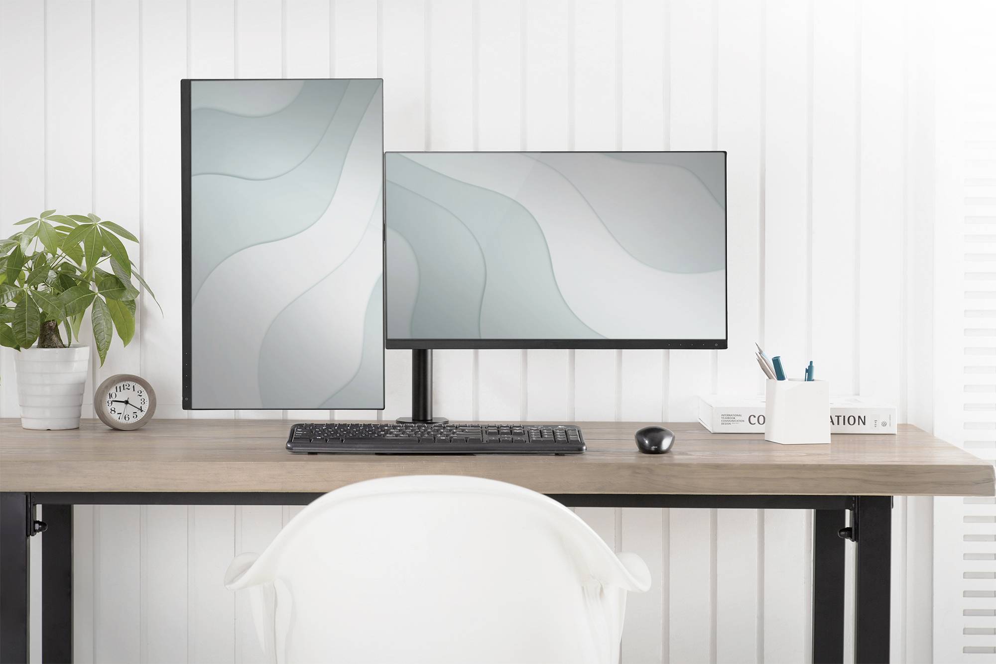 Ein moderner, aufgeräumter Schreibtisch mit zwei Monitoren, einer vertikal, einer horizontal. Links steht eine Pflanze, rechts Schreibutensilien.
