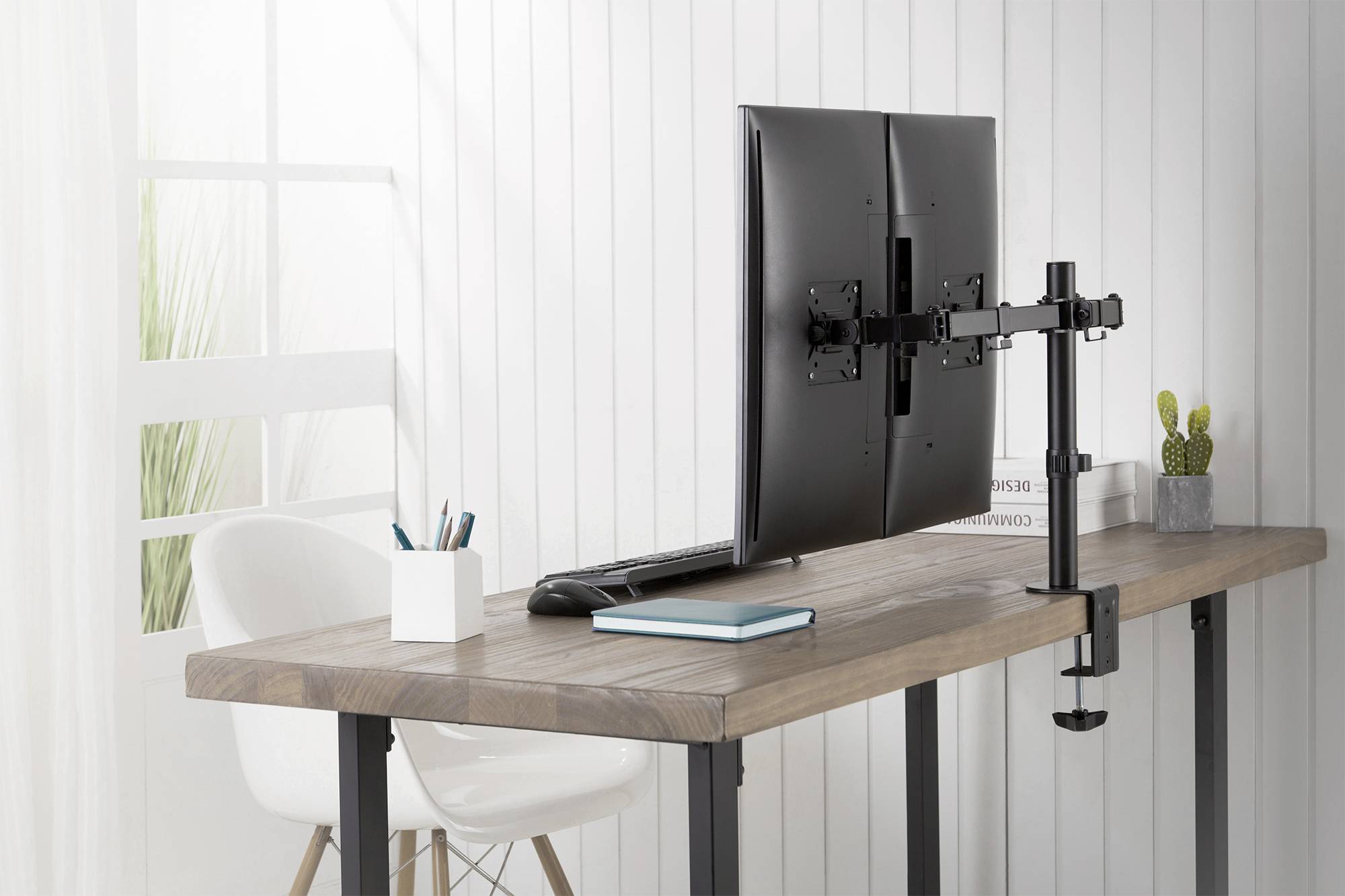 Ein Schreibtisch mit einem doppelt montierten Monitorarm, zwei Monitoren, Laptop, Notizbüchern und Büromaterial in einem hellen Raum.