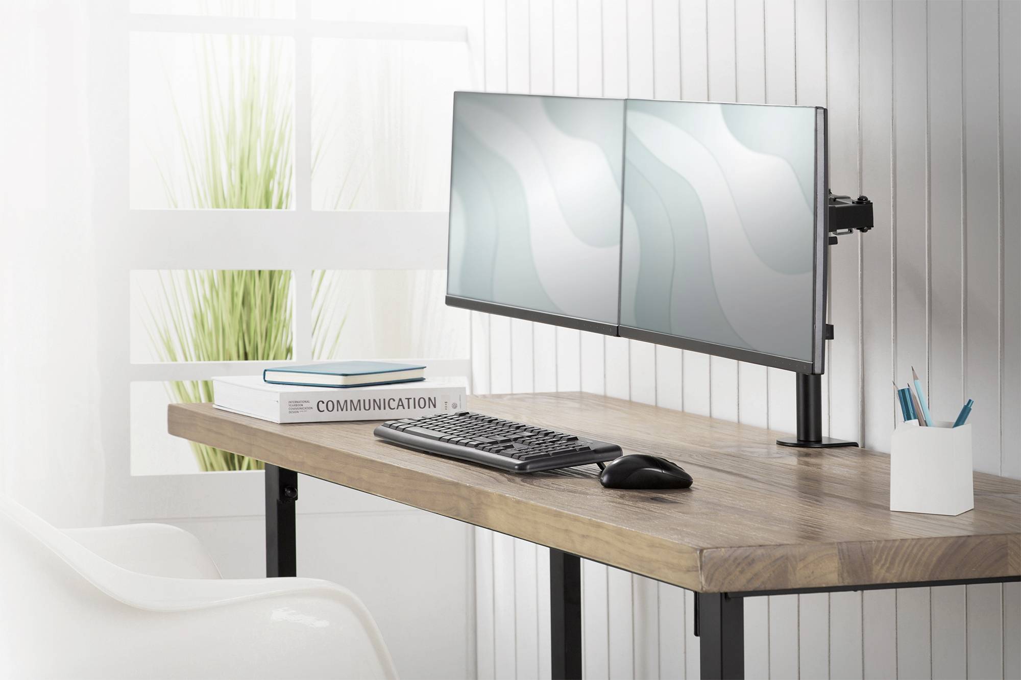 Ein Schreibtisch mit einem Doppelmonitor-Setup, daneben eine Tastatur und Maus. Ein Buch mit dem Titel 'Communication' liegt links.