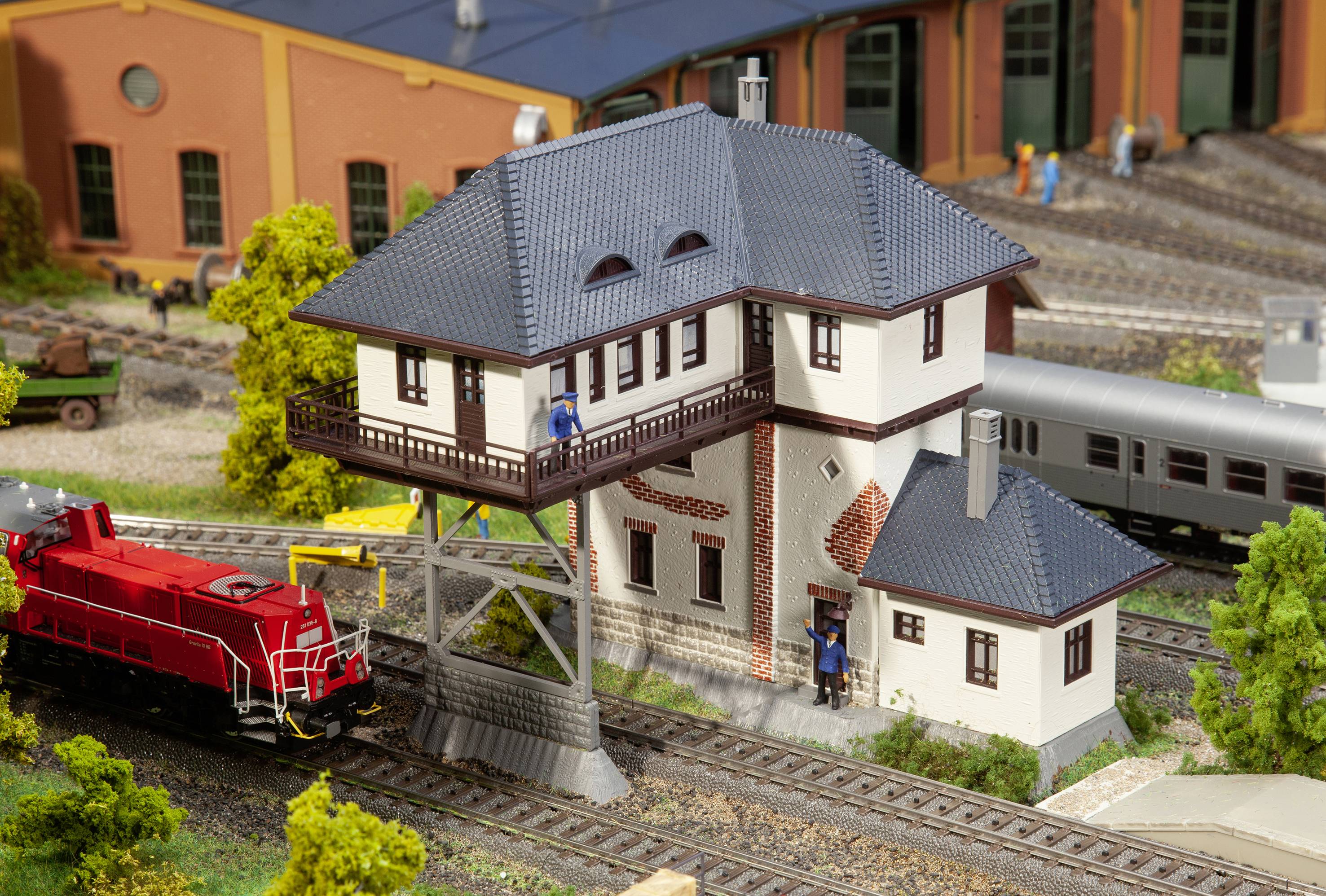 Ein Miniaturmodell zeigt ein Eisenbahndepot mit einem roten Zug, einem weißen Gebäude und mehreren Figuren.