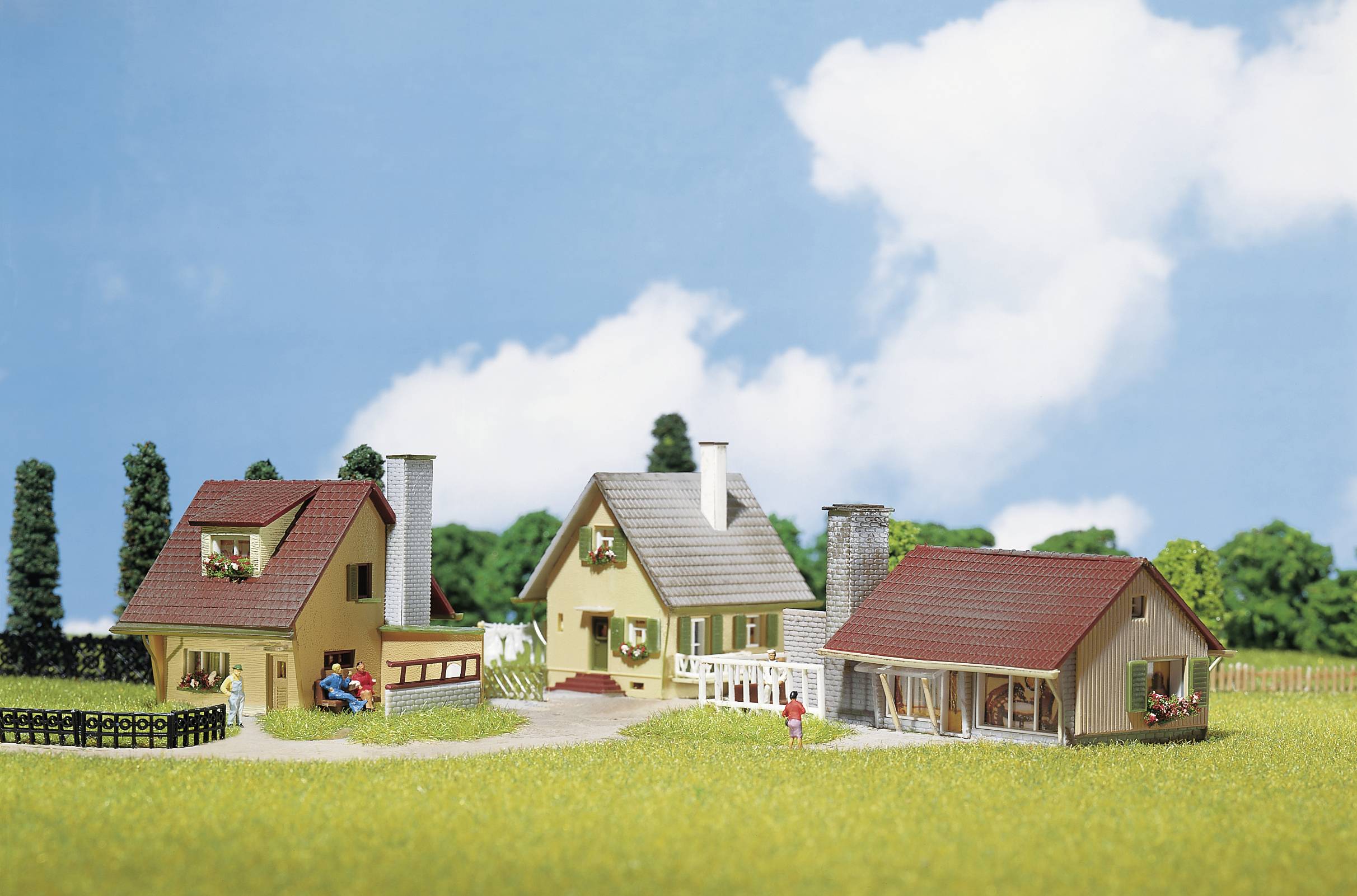 Drei Modellhäuser in einer ländlichen Umgebung, umgeben von Bäumen und Wiese, unter blauem Himmel mit einigen weißen Wolken.