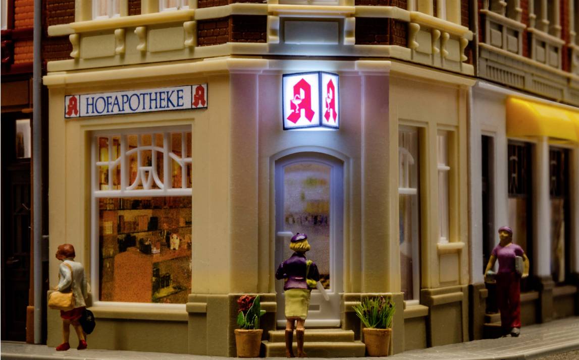 Eine Miniaturapotheke an einer Straßenecke, beleuchtet bei Nacht, mit einem leuchtenden Apothekenschild und Figuren in Bewegung.