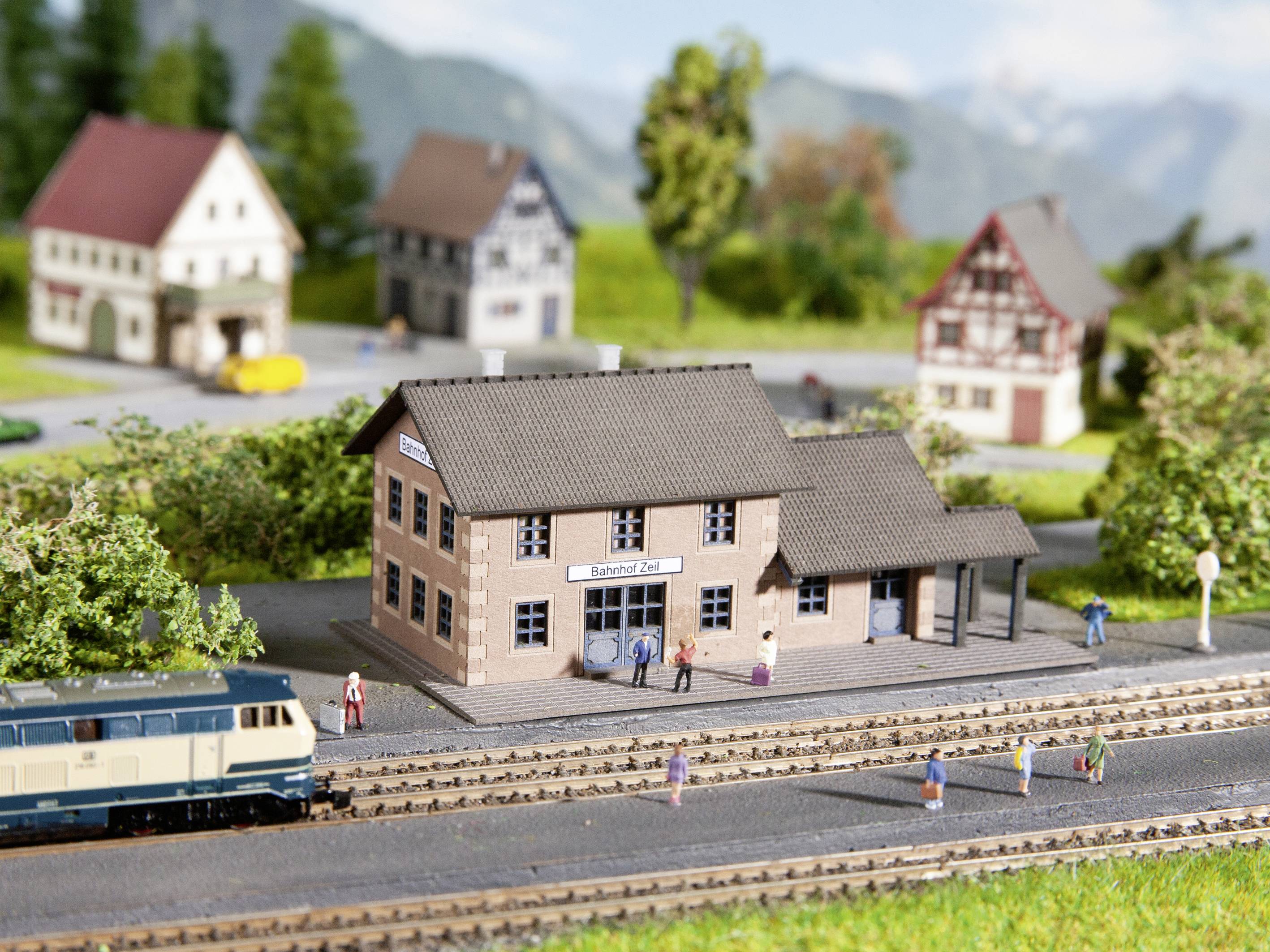 Eine Miniatureisenbahnlandschaft zeigt ein ländliches Bahnhofsgebäude mit vorbeifahrendem Zug, umgeben von verkleinerten Häusern und Bäumen.