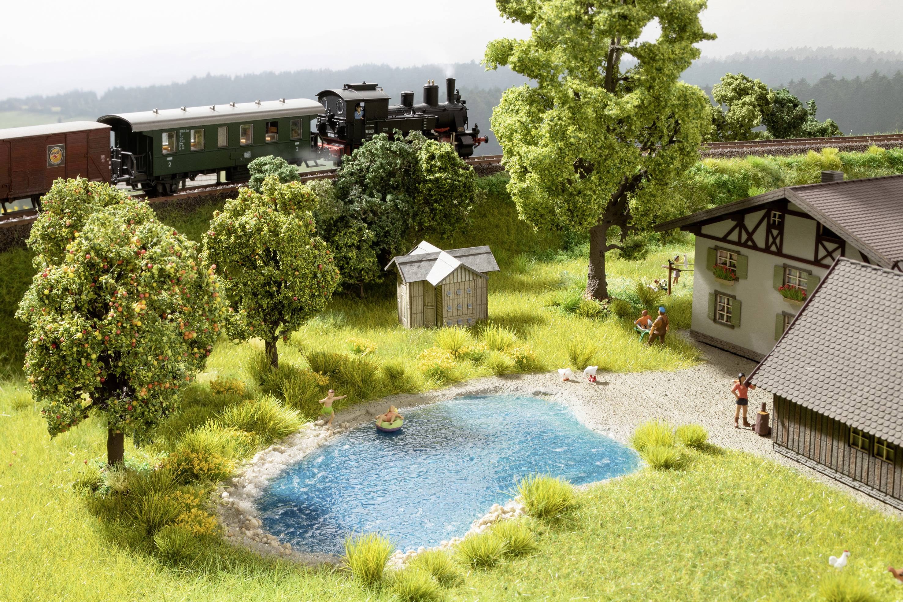 Ein Modellzug fährt über ein Gleis hinter einer Miniaturlandschaft mit Teich, umgeben von Bäumen und einem kleinen Gebäude.