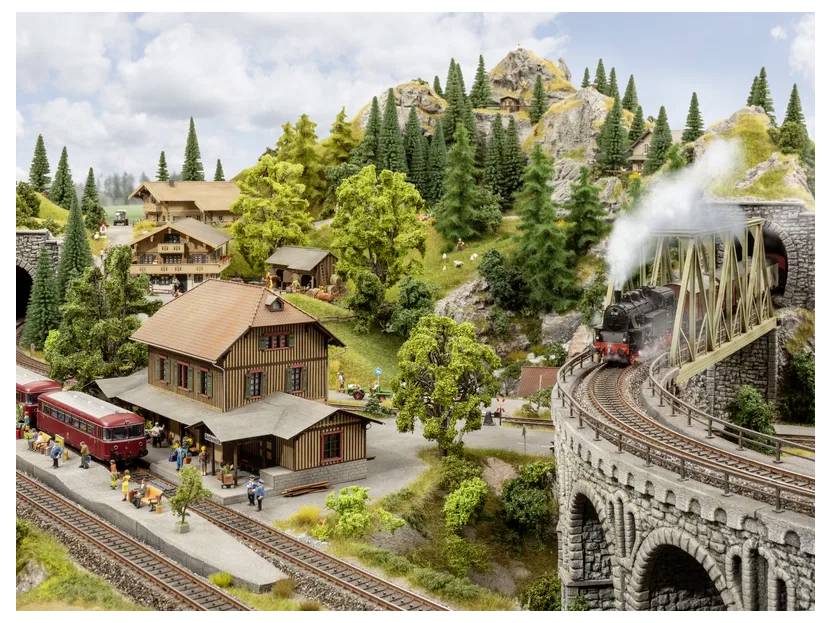 A detailed model train set depicting a scenic mountain village with a station, red trains, lush trees, rocky hills, and a bridge.