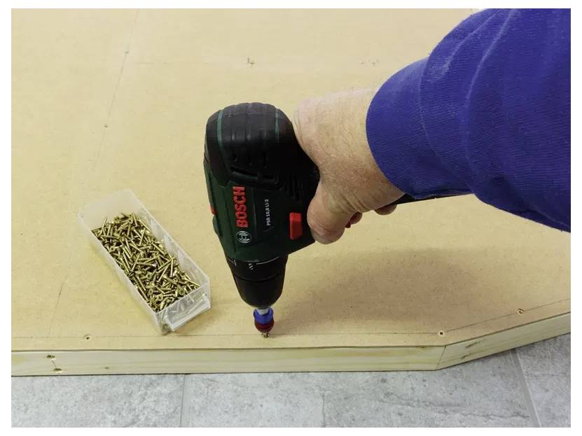 A hand uses a Bosch electric screwdriver to fasten screws into plywood. A box of screws lies nearby.