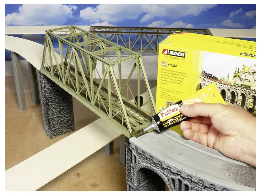 A person applies adhesive to a model bridge component, with a Noch brand product box visible. The scene shows a model bridge setup.