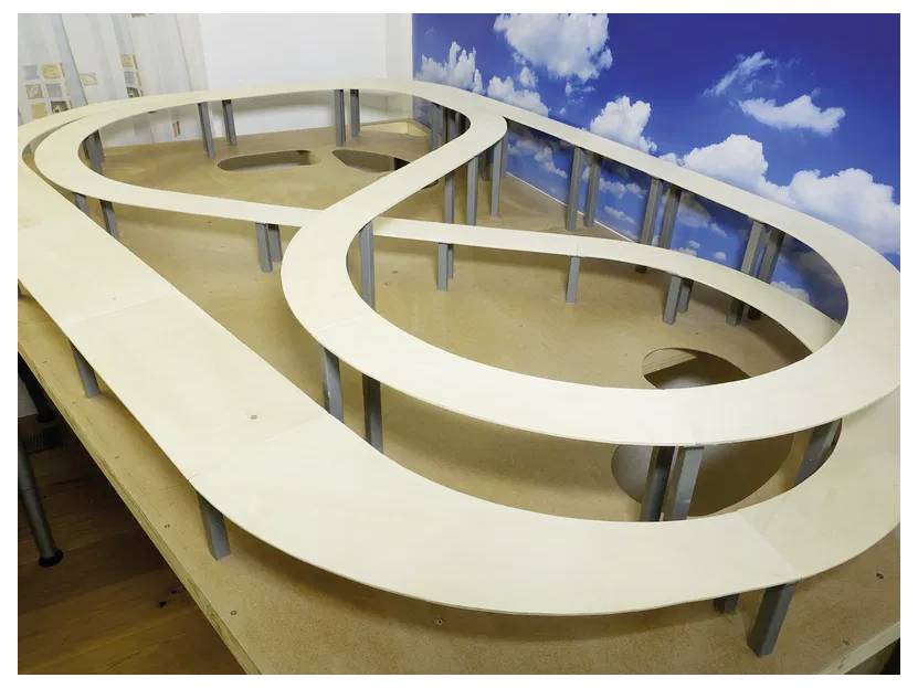 Model race track on a wooden table with multiple elevated loops, set against a backdrop of a sky with clouds.
