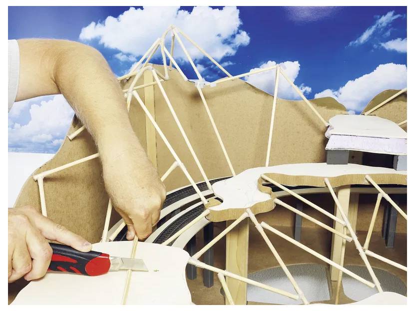 A person is constructing a model structure using sticks and cardboard, with a blue sky backdrop.