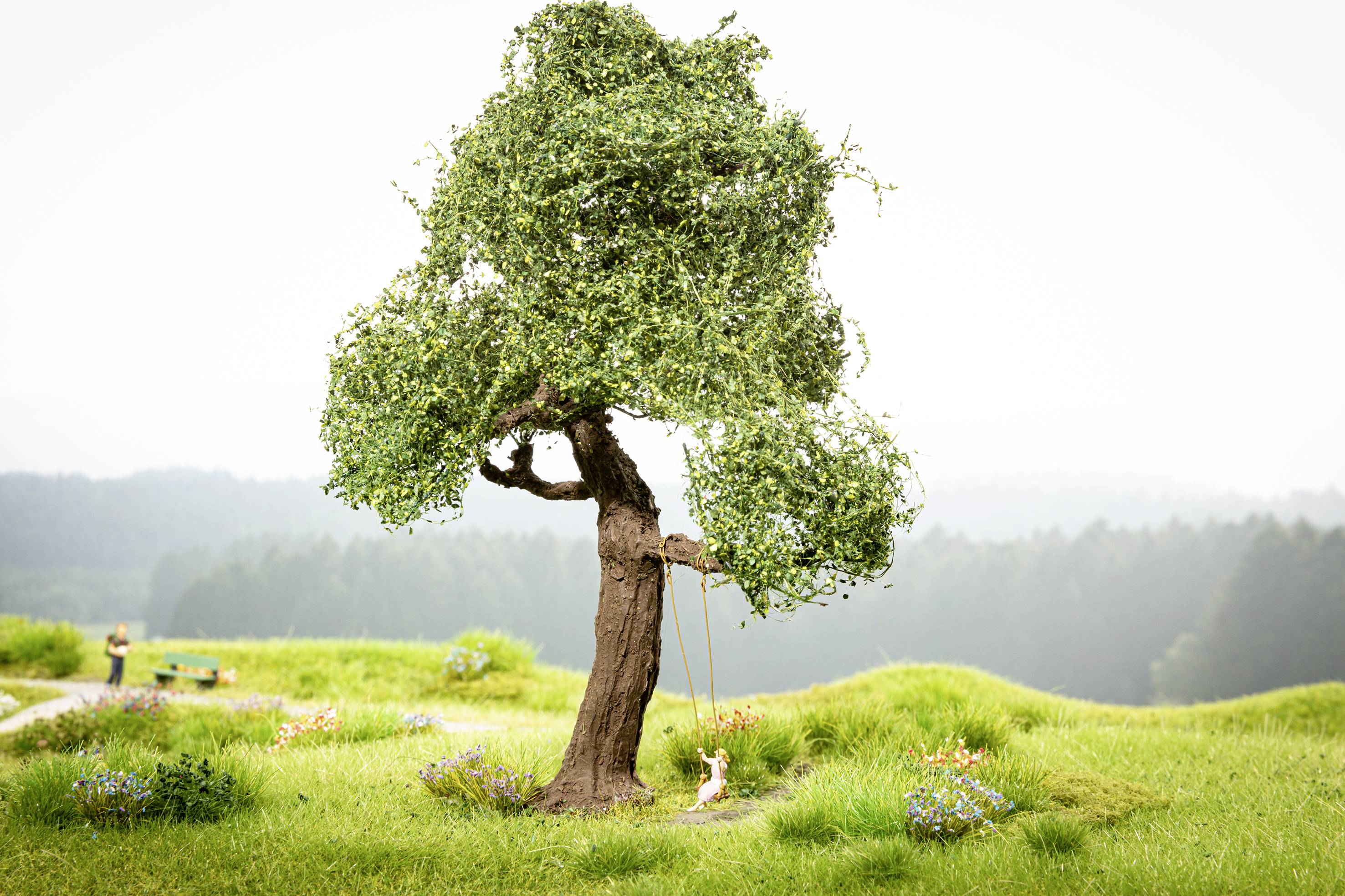 Eine Miniaturbaumlandschaft zeigt einen Baum mit Schaukel und einem kleinen Hasen darunter, umgeben von grüner Wiese und Blumen.