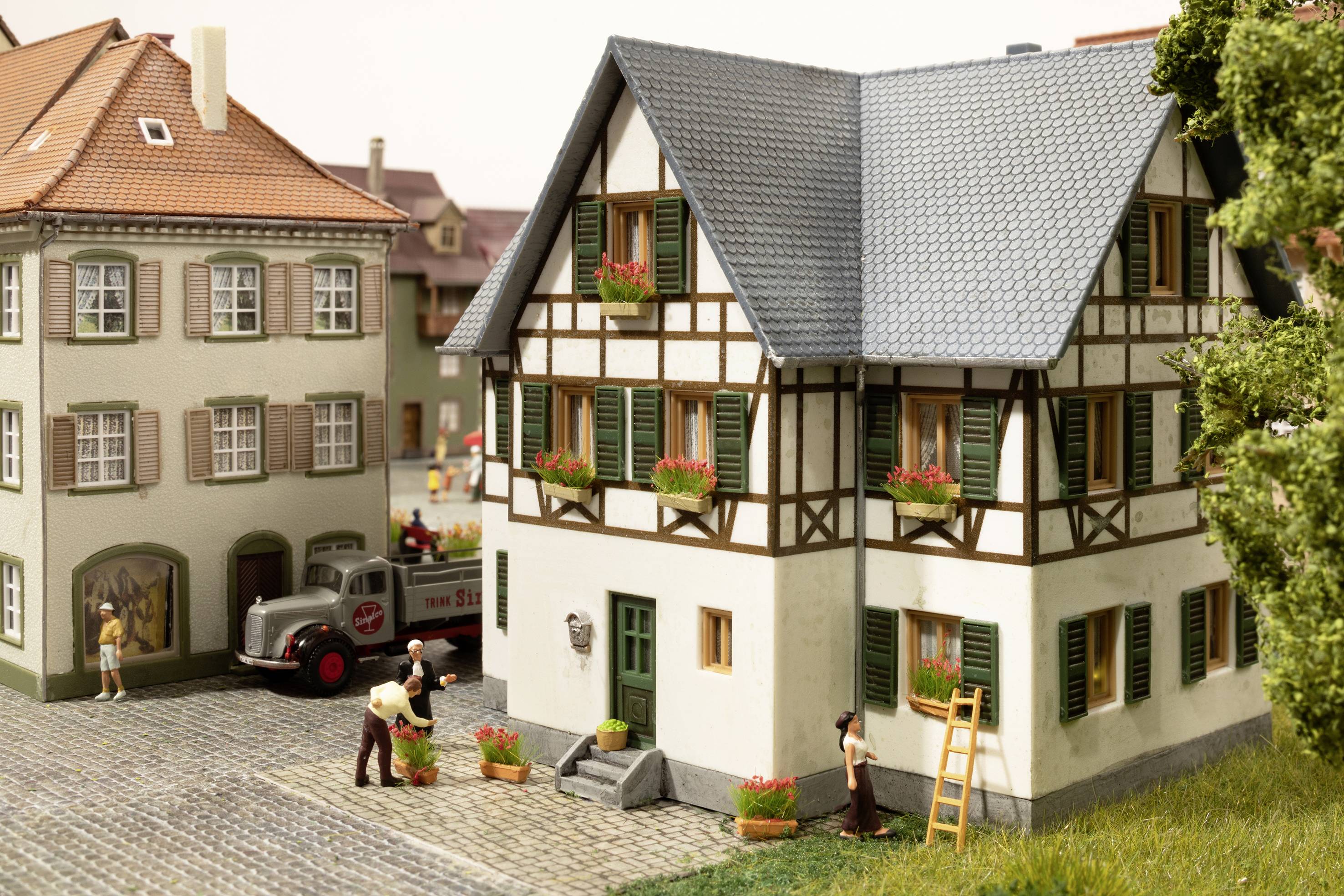 Miniatur eines Fachwerkhauses mit blühenden Blumenkästen, umgeben von Figuren in einem Dorf. Ein Lieferwagen parkt daneben.