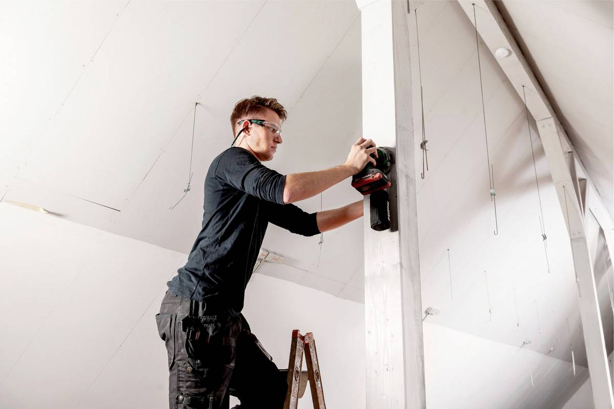 Eine Person arbeitet in einem Dachboden auf einer Leiter mit einer Schleifmaschine an einem Holzpfeiler und trägt Schutzbrille.