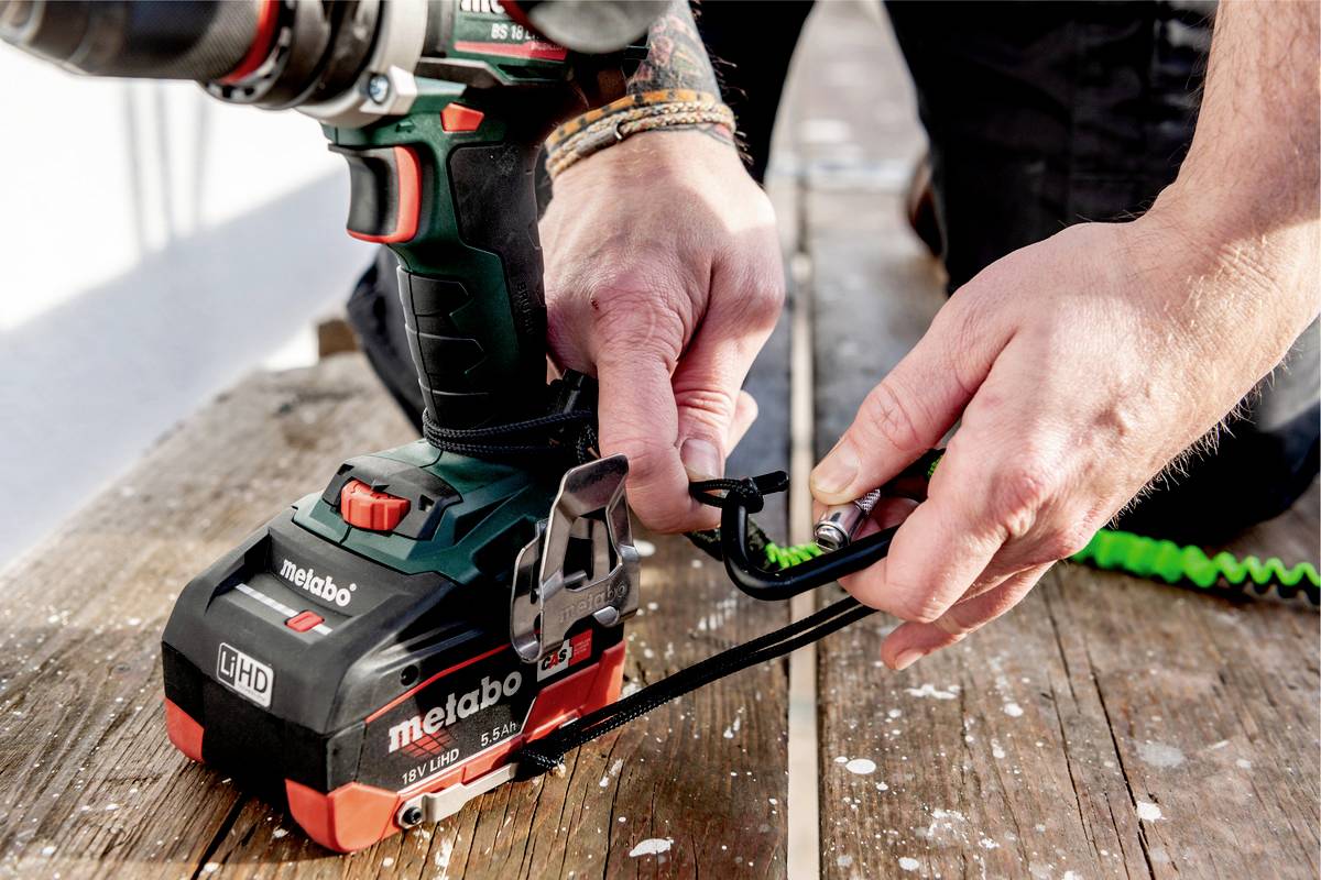 Eine Person befestigt ein Sicherheitsseil an einer Bohrmaschine auf einem Holzboden. Die Bohrmaschine ist von der Marke Metabo.
