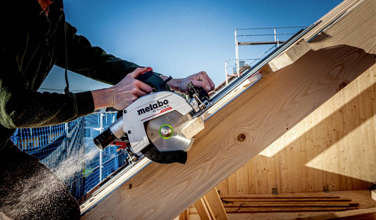 Eine Person verwendet eine Handkreissäge, um einen Holzbalken auf einer Baustelle zu schneiden. Heller Himmel und Baugerüst im Hintergrund.