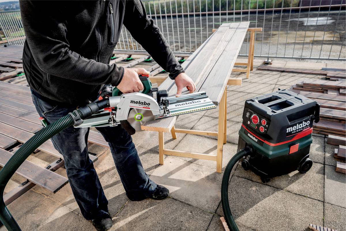 Person arbeitet mit einer Metabo Kappsäge auf einer Terrasse. Ein Staubsauger ist zur Staubabsaugung angeschlossen. Umgebung wirkt industriell.