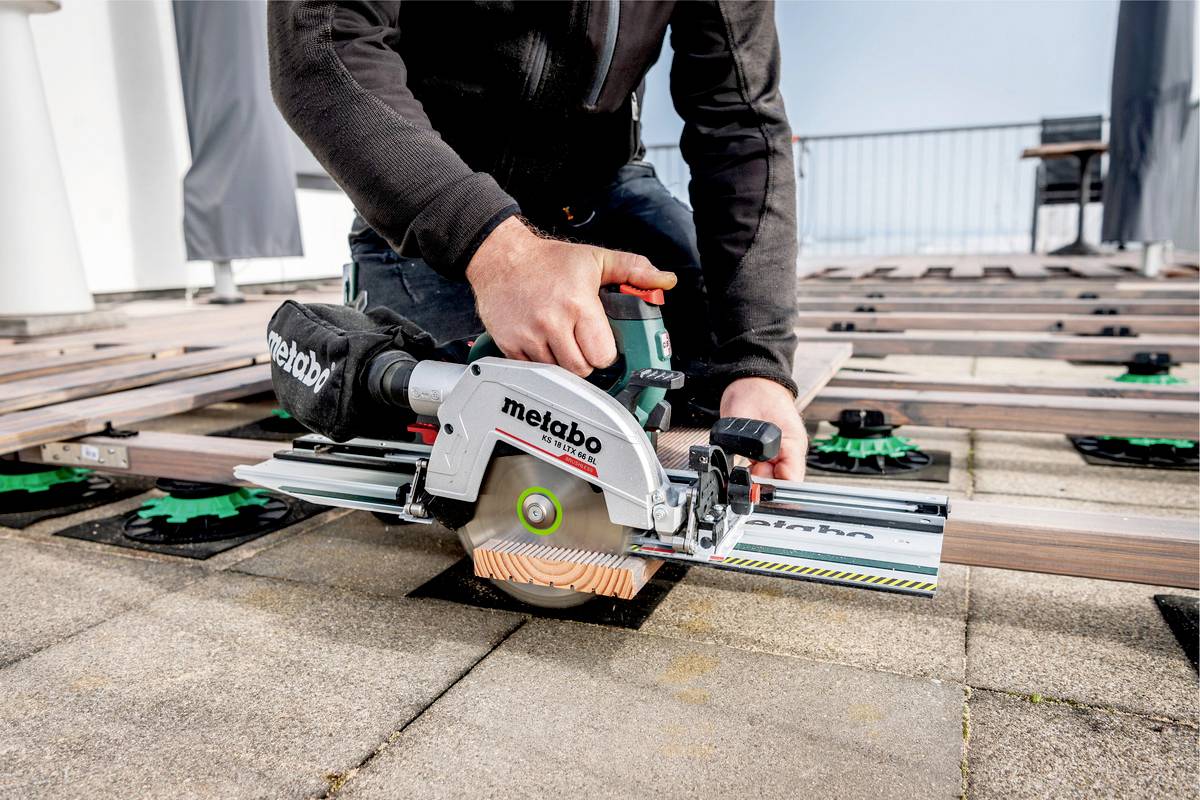 Eine Person sägt Holzbretter auf einer Terrasse mit einer Metabo-Handkreissäge. Im Hintergrund sind weitere Bretter und eine Balustrade sichtbar.