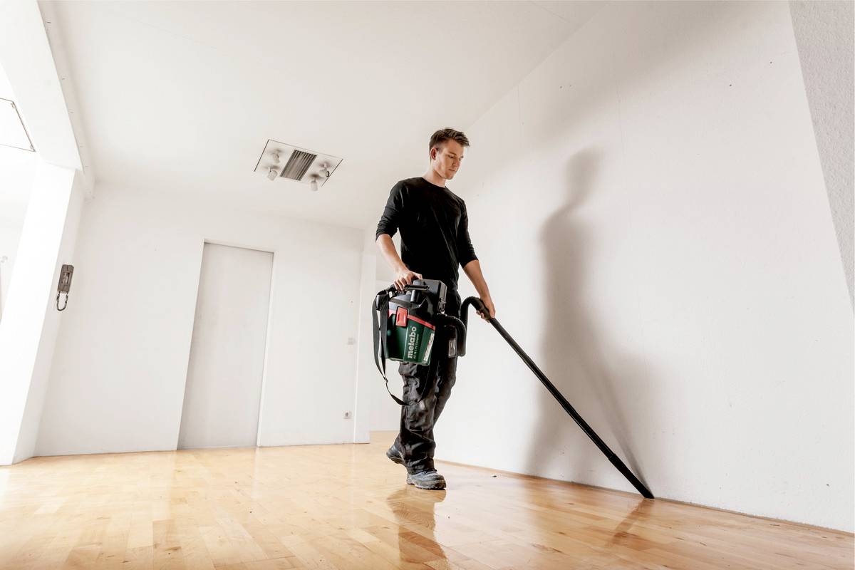 Eine Person saugt mit einem Staubsauger einen leeren Raum mit Holzboden. Die Wände sind weiß, der Raum ist hell erleuchtet.